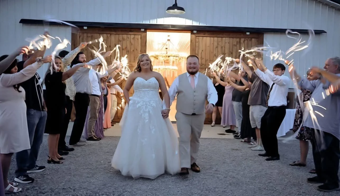 A bride and groom walking hand in hand through a tunnel of guests holding sparklers during a wedding celebration outside a barn.