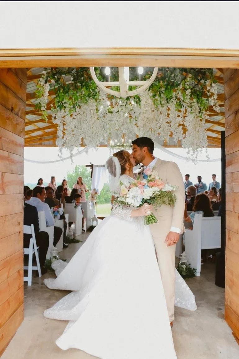 Bride and groom kiss during their wedding ceremony under floral decorations, with guests seated nearby and a view of greenery outside.
