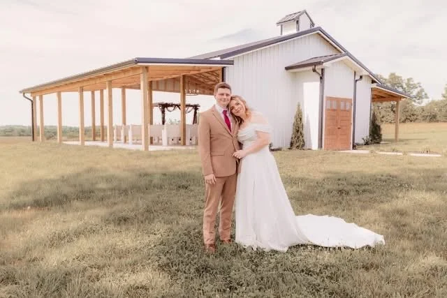 A newlywed couple in wedding attire standing on a grassy field in front of a white barn with a porch and a small cupola on top.