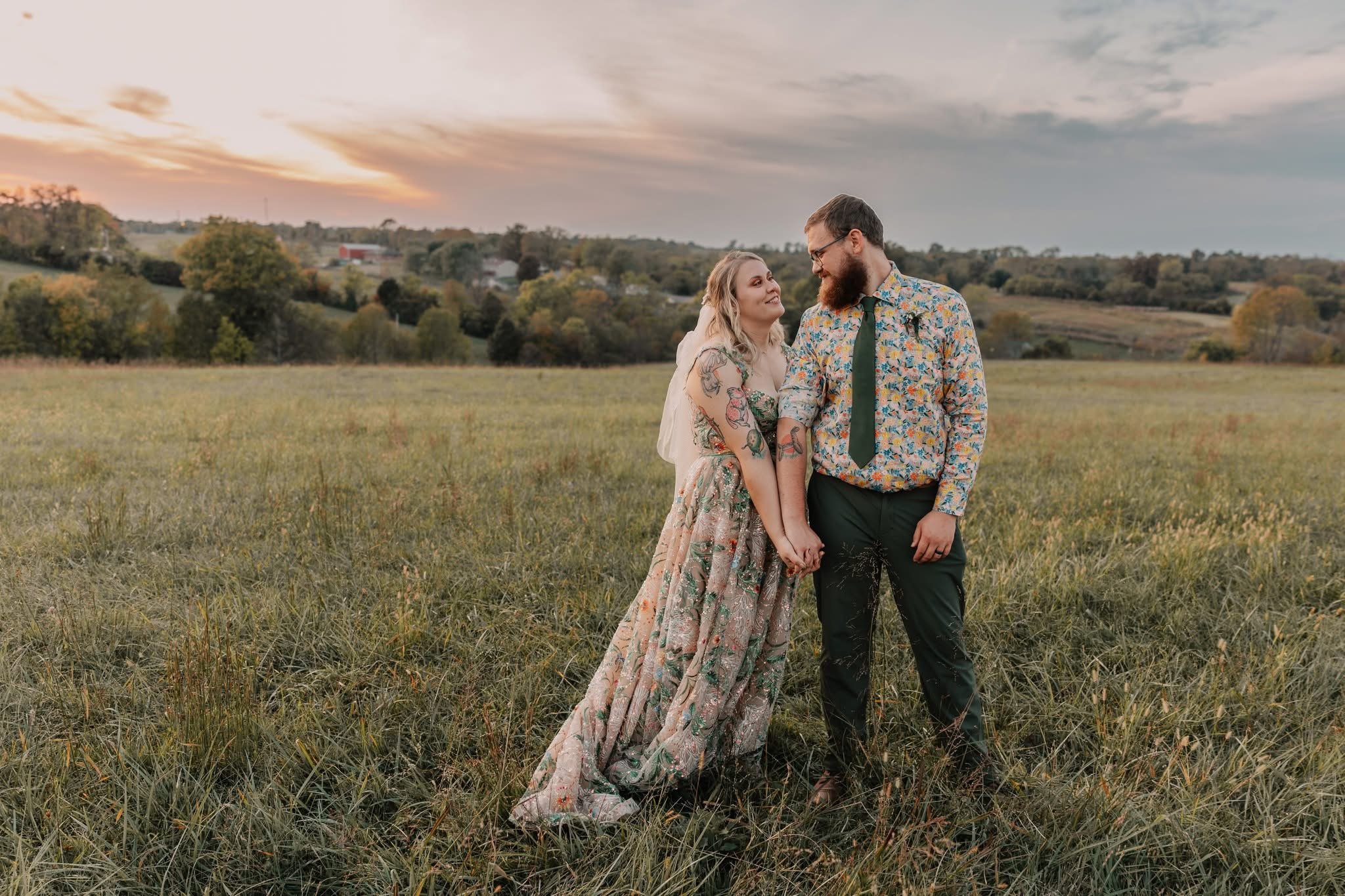 A couple standing hand in hand in a grassy field at sunset, smiling and looking at each other. The woman has tattoos on her arms and is wearing a floral dress. The man has a beard, glasses, and is dressed in a floral shirt with a tie. The background shows rolling hills, trees, and a cloudy sky with the sun setting.