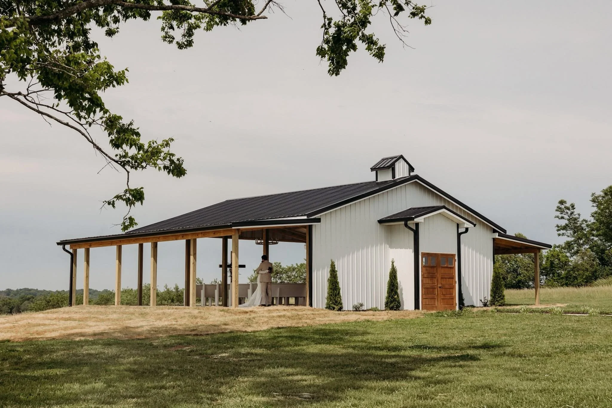 An outdoor house under construction, with a partially finished white exterior and a metal roof. There is a covered porch area with a person dressed in white standing underneath it. The background shows a cloudy sky and green trees.