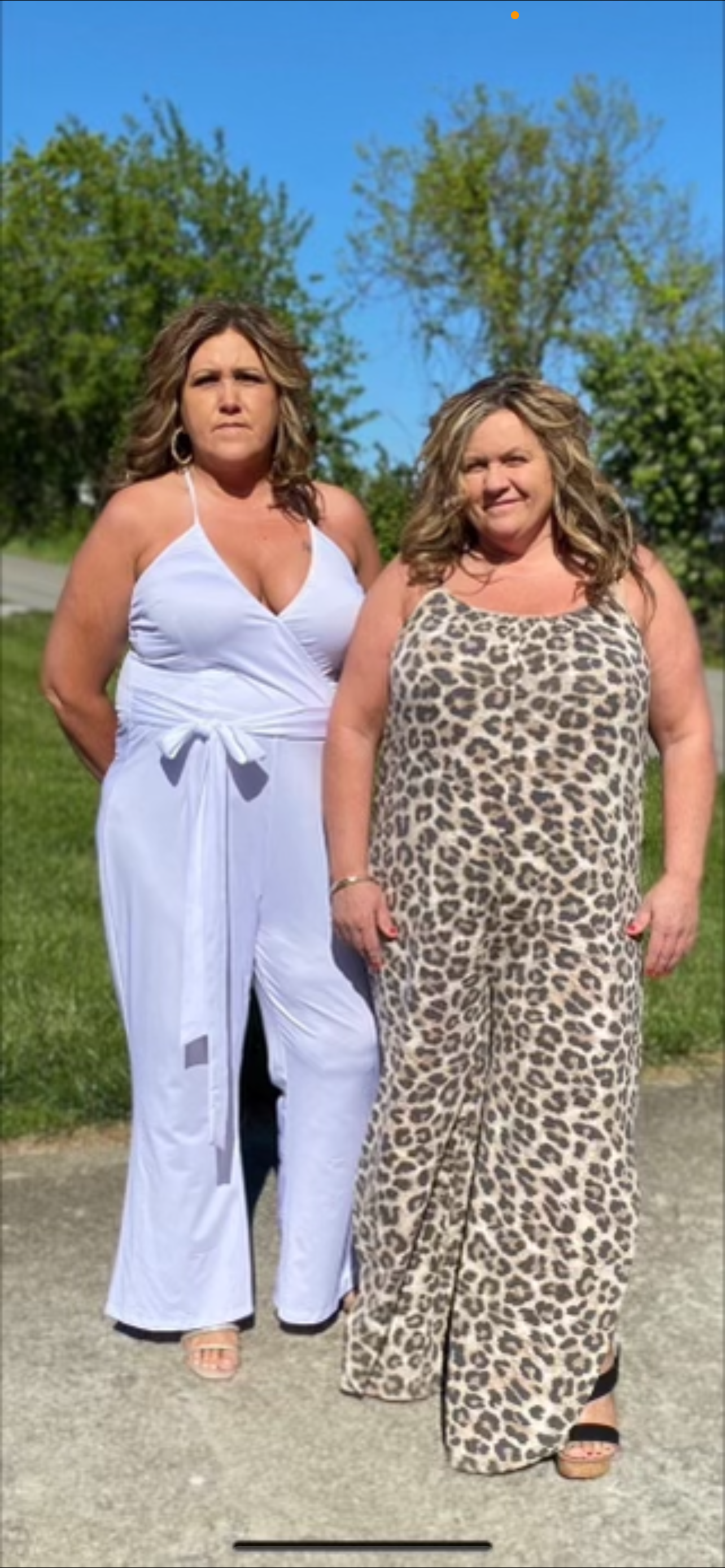 Two women standing outdoors on a sunny day, one wearing a white sleeveless jumpsuit and the other wearing a leopard print dress, with green trees and a blue sky in the background.
