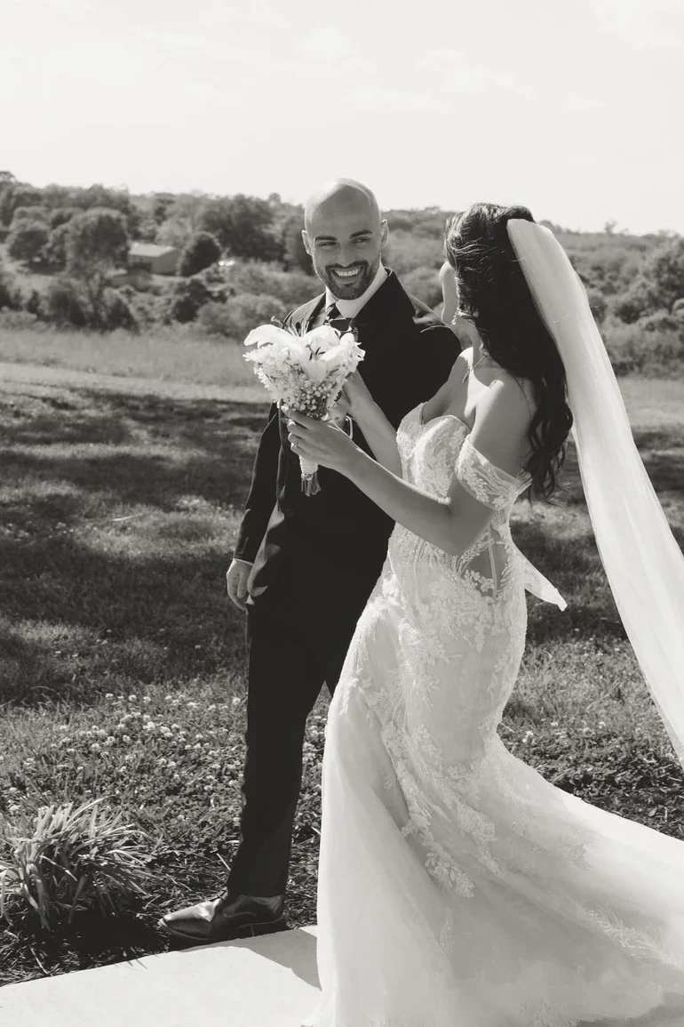A black and white photo of a bride and groom outdoors; the groom is smiling and holding a bouquet, the bride is wearing a lace wedding dress with a veil.