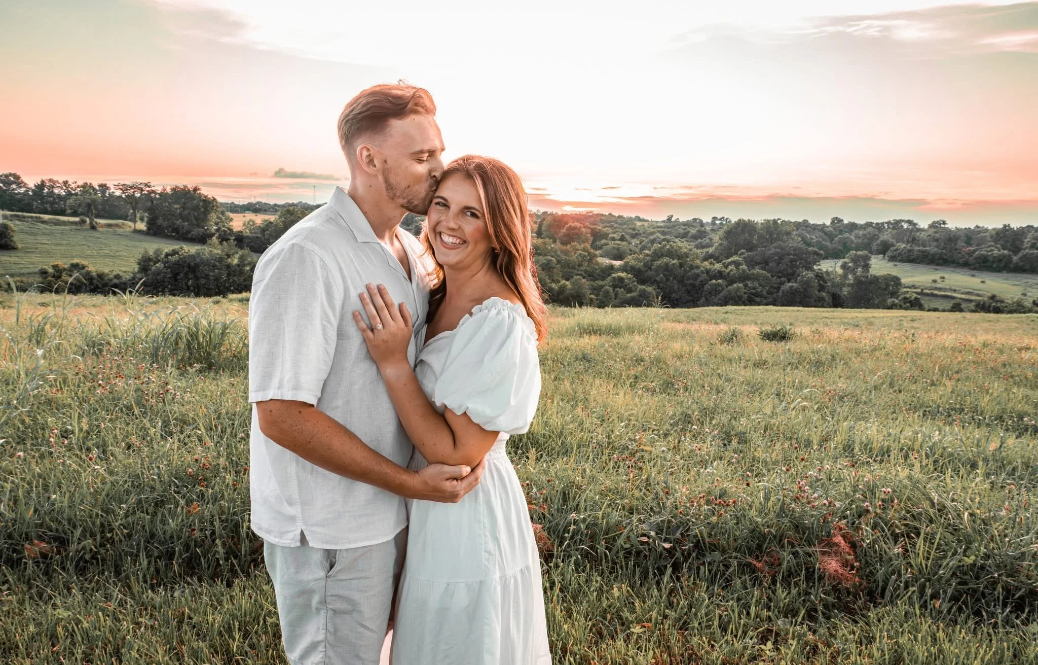 A couple standing in a field during sunset, with the man kissing the woman on the forehead, both smiling and dressed in white, surrounded by greenery and rolling hills.