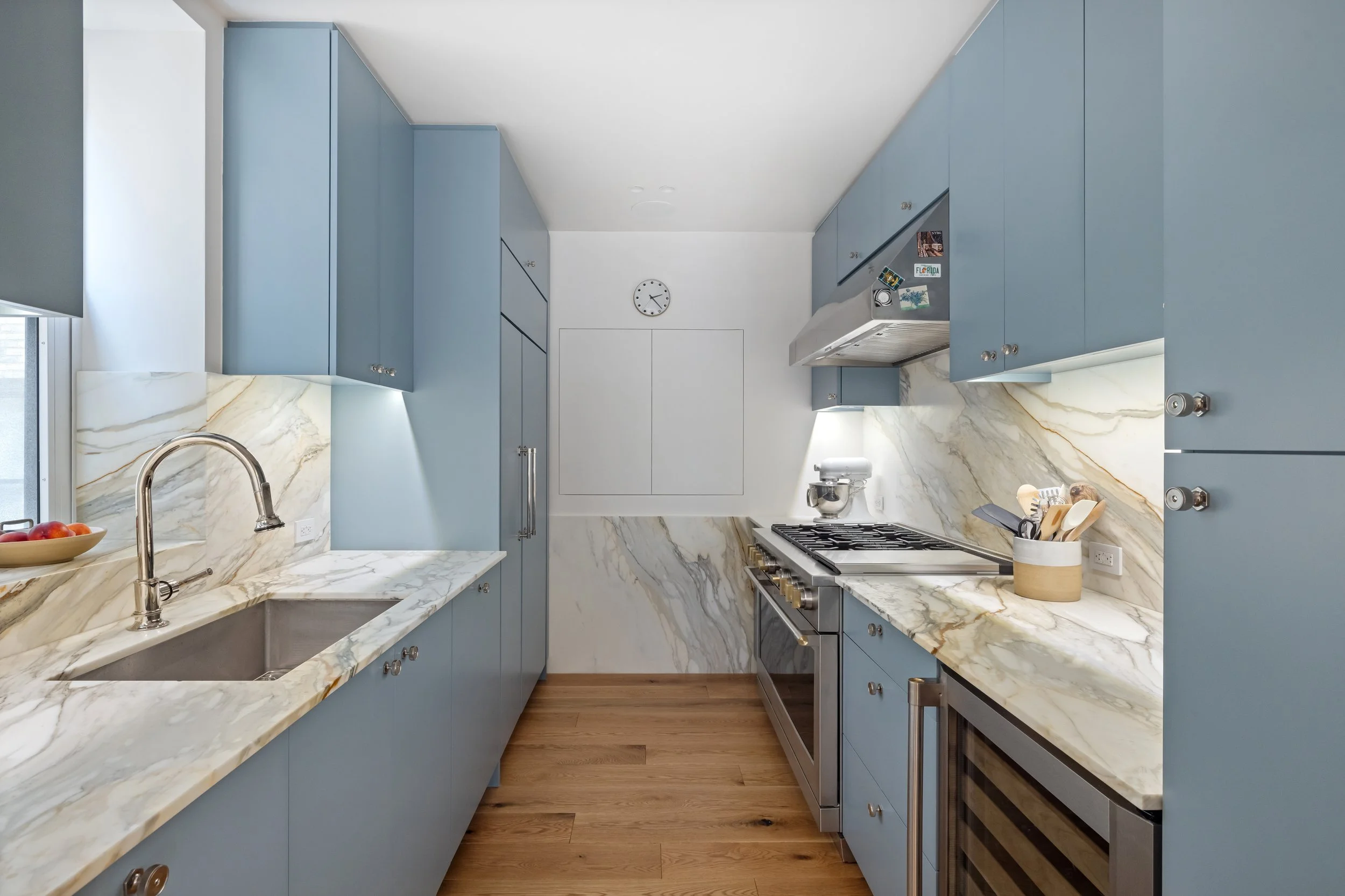 A modern kitchen with blue cabinets, white marble countertops, a stainless steel stove, a stainless steel range hood with magnets, a wall clock, a window with a roller blind, a bowl of apples, and kitchen utensils.