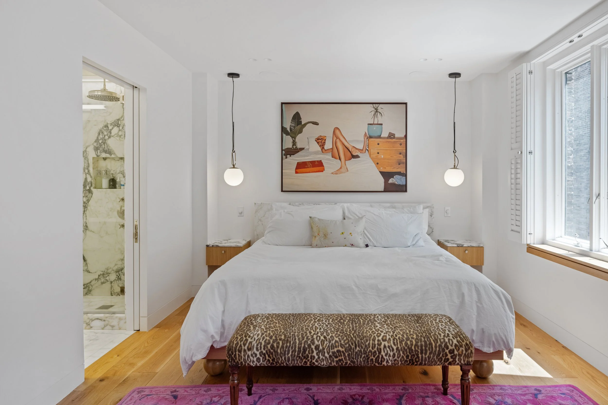 A bedroom with a white bed, a framed art piece above the bed, two hanging pendant lights, a leopard print ottoman at the foot of the bed, and a pink rug. To the left, there is a doorway to a bathroom with a marble shower.