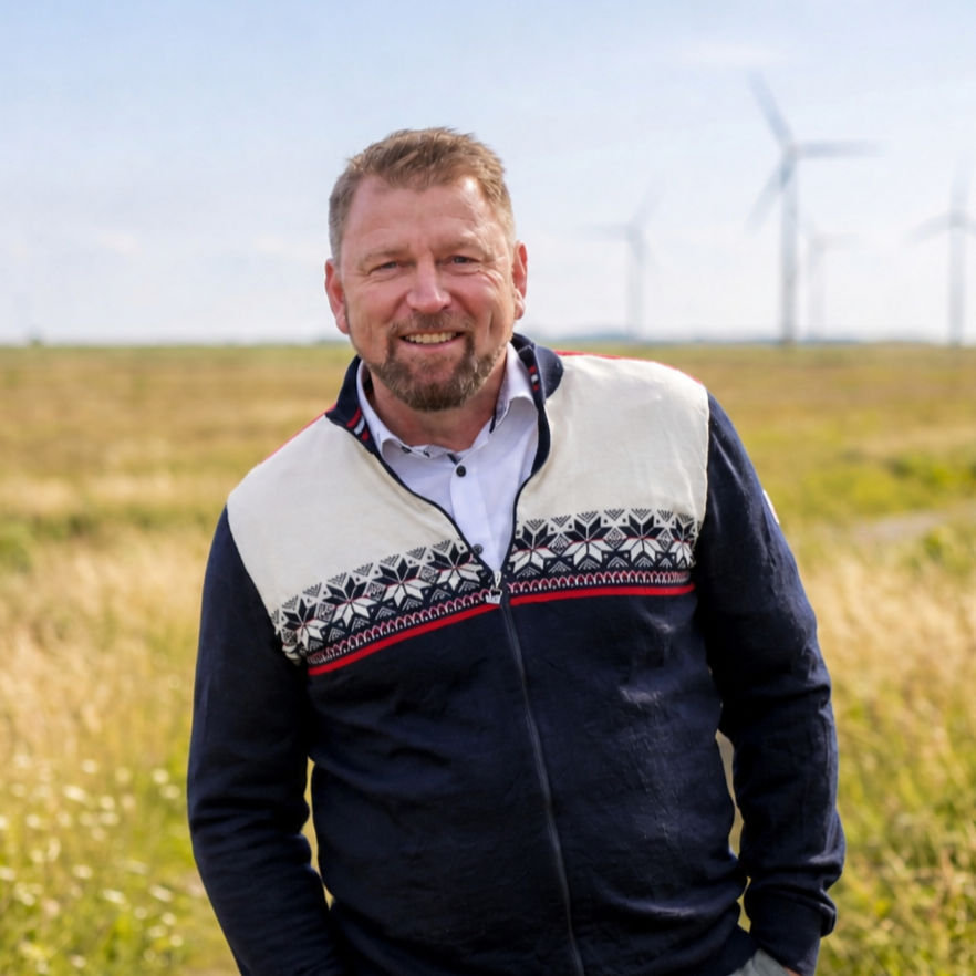 Ein Mann steht in einem Feld mit Windturbinen im Hintergrund, lächelt und trägt eine schwarze Jacke mit weißem, rotem und gemustertem oberen Teil.