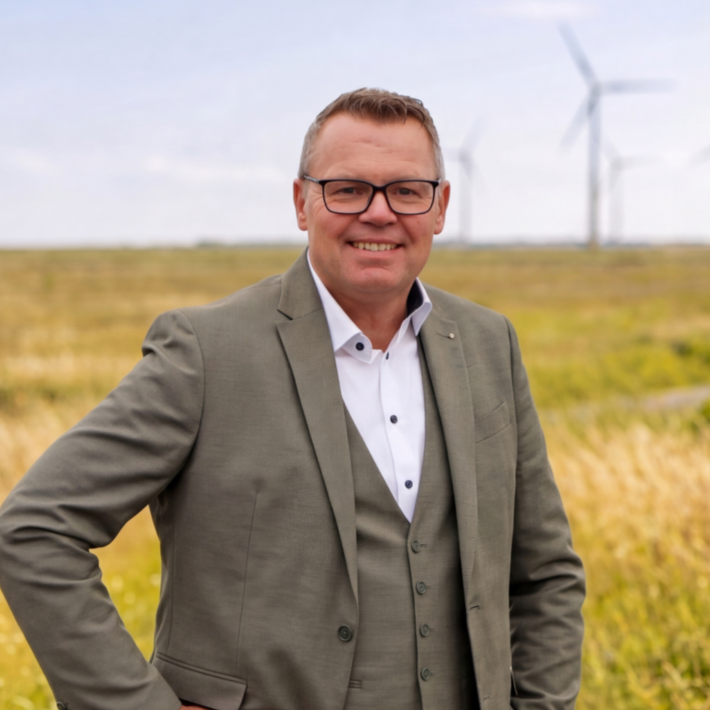Ein Mann trägt einen grauen Anzug und steht vor Windrädern auf einer Wiese, Himmel ist bewölkt.