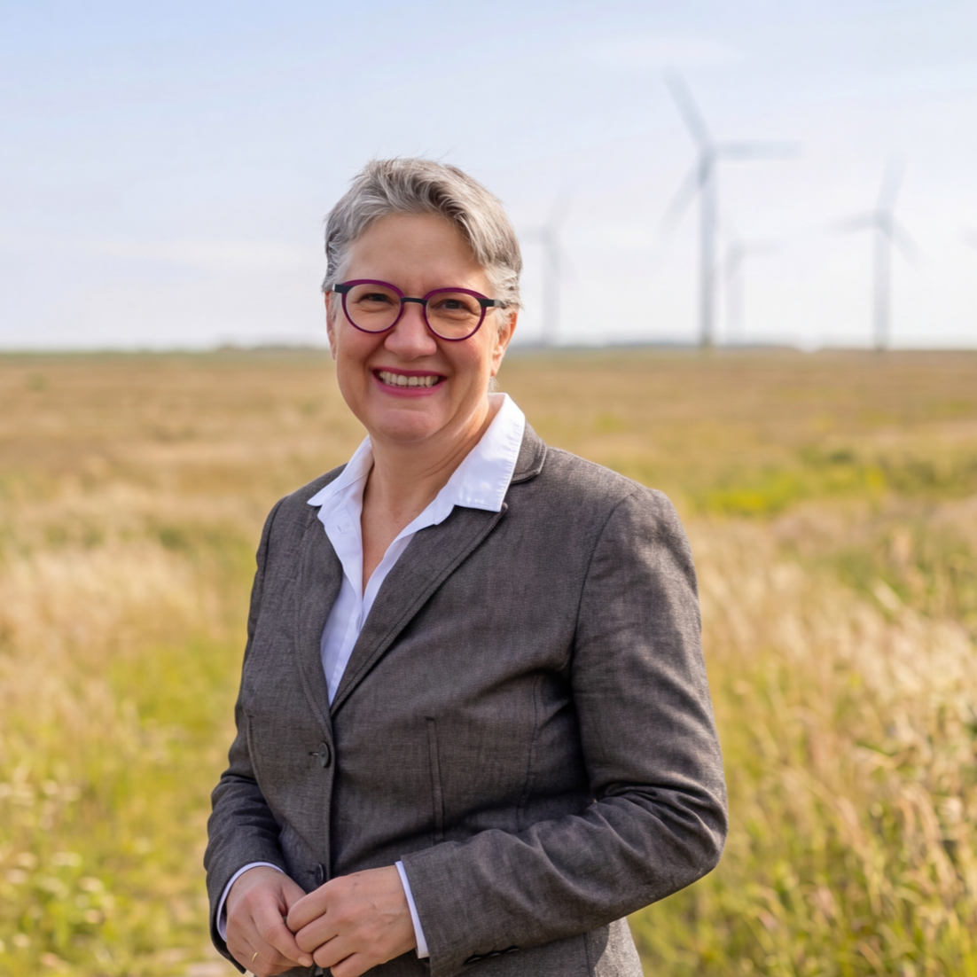 Eine lächelnde Frau in einem grauen Geschäftsanzug steht in einem offenen Feld mit Windturbinen im Hintergrund.