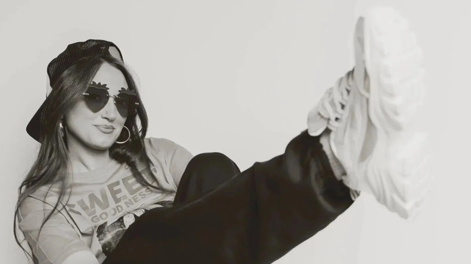 A woman with long hair wearing sunglasses, a cap, and a T-shirt with the word 'SWEETNESS' on it, sitting against a plain wall, with her legs extended and her foot pointed towards the camera.