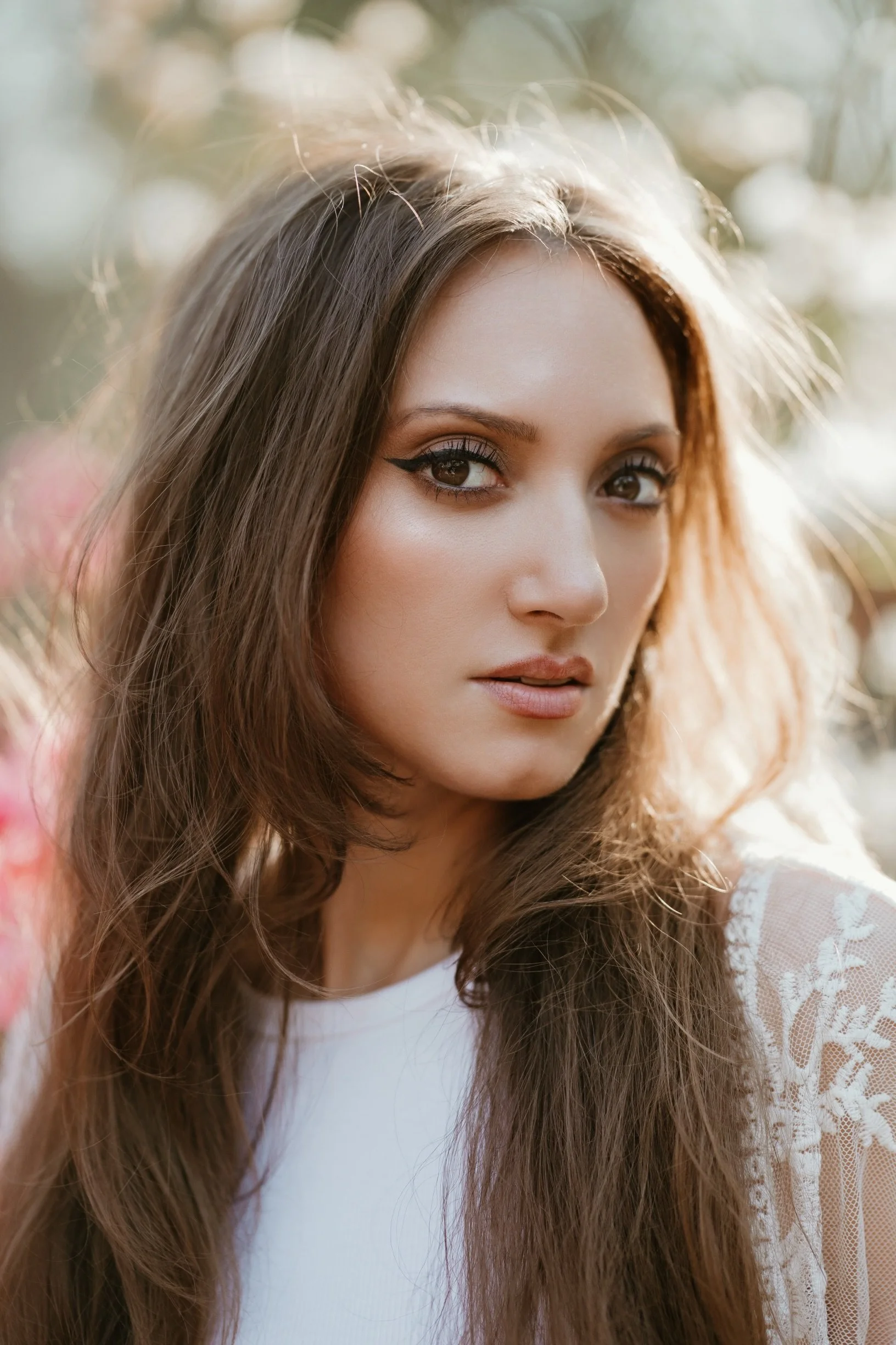 A close-up of a young woman with long, wavy brown hair and striking eye makeup, standing outdoors with sunlight illuminating her hair.