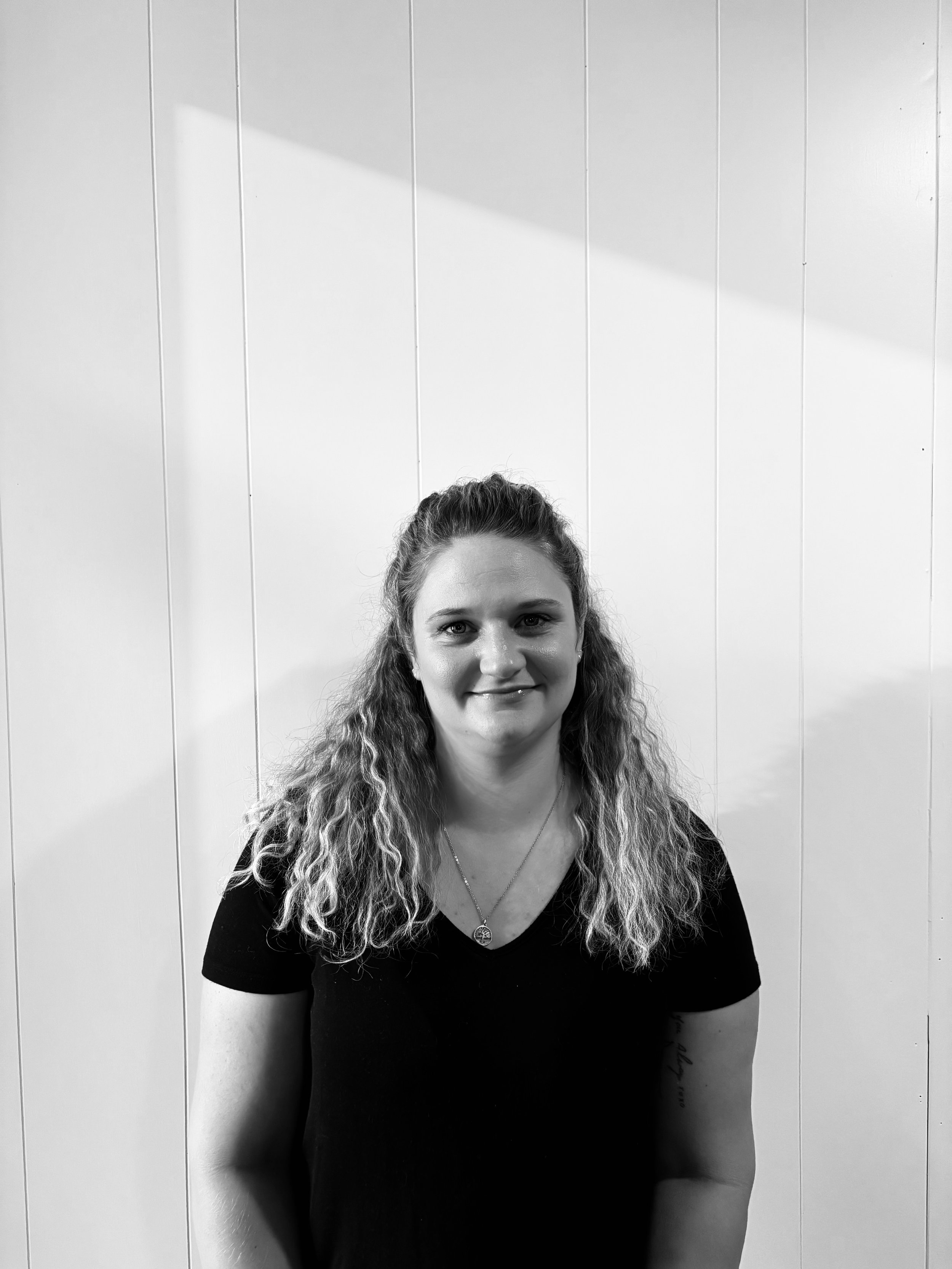 A young woman with curly hair and a necklace smiling in front of a plain wall.