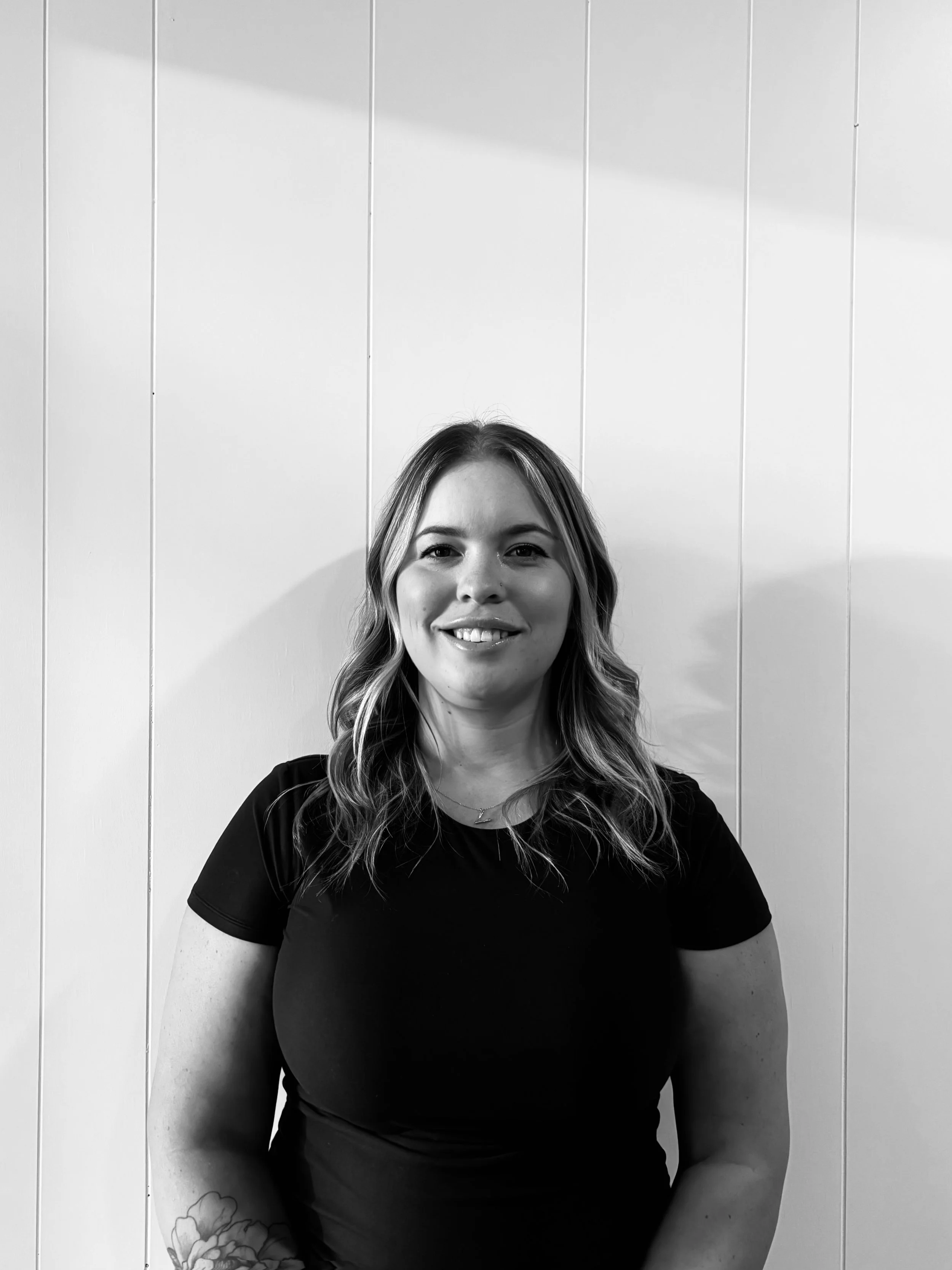 Black and white photo of a smiling woman with wavy hair, wearing a dark t-shirt, standing against a paneled wall.