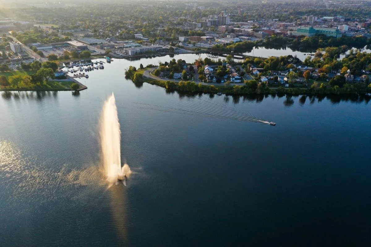 Aerial view of a city with a large body of water, a fountain spraying water into the air, a speedboat creating ripples, and residential houses surrounded by trees.
