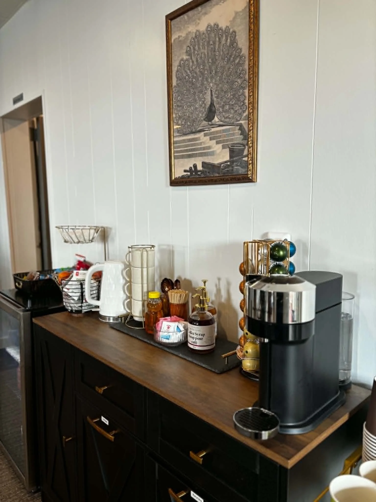 A kitchen coffee station with a wooden counter, coffee machine, and various coffee supplies including syrups, cups, and a basket of tea bags. A framed picture of a peacock is hanging on the wall above the counter.