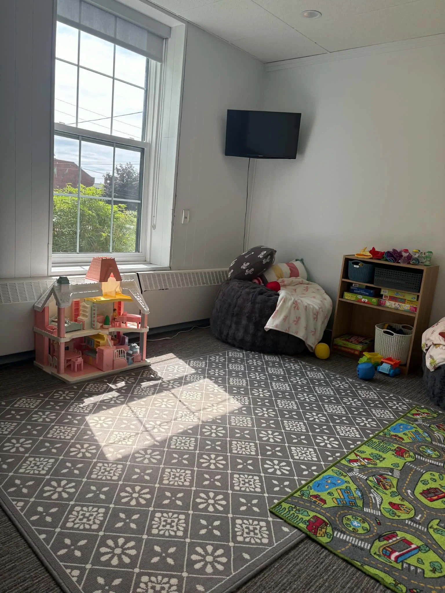 A room with a large window, a television mounted on the wall, a small bookshelf with toys and books, a dollhouse on the floor, a blanket-covered chair, and a play mat with a city street design.