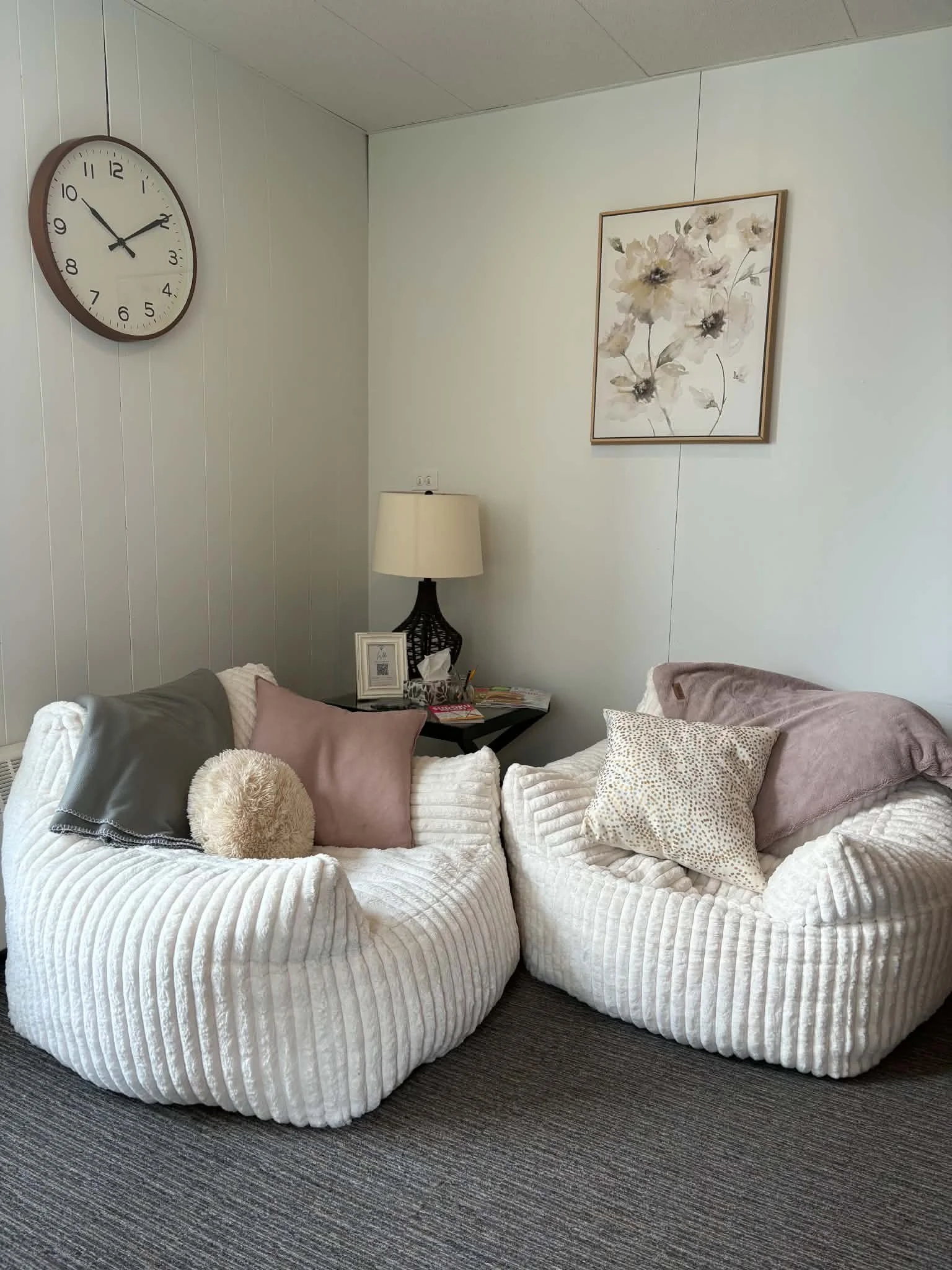 Two plush white sofas with various pillows and a round beige furry pillow, situated on a carpeted floor in a living room with a wall clock, framed flower art, a table with magazines, a lamp, and a small picture frame.