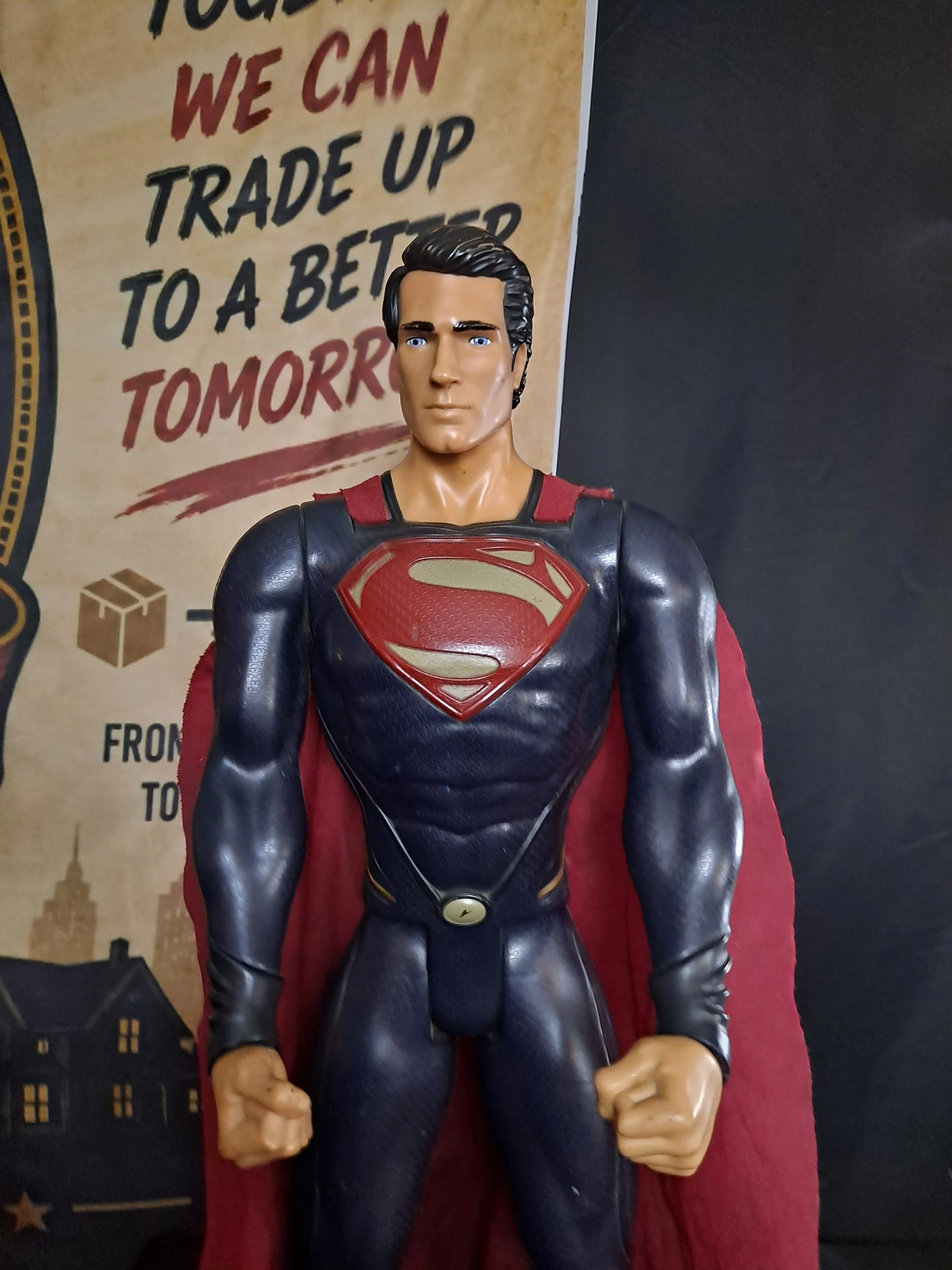 Figure of Superman in black costume with red cape, fists clenched at sides, standing in front of a poster with the words 'Trade up to a better tomorrow' and cityscape illustration.