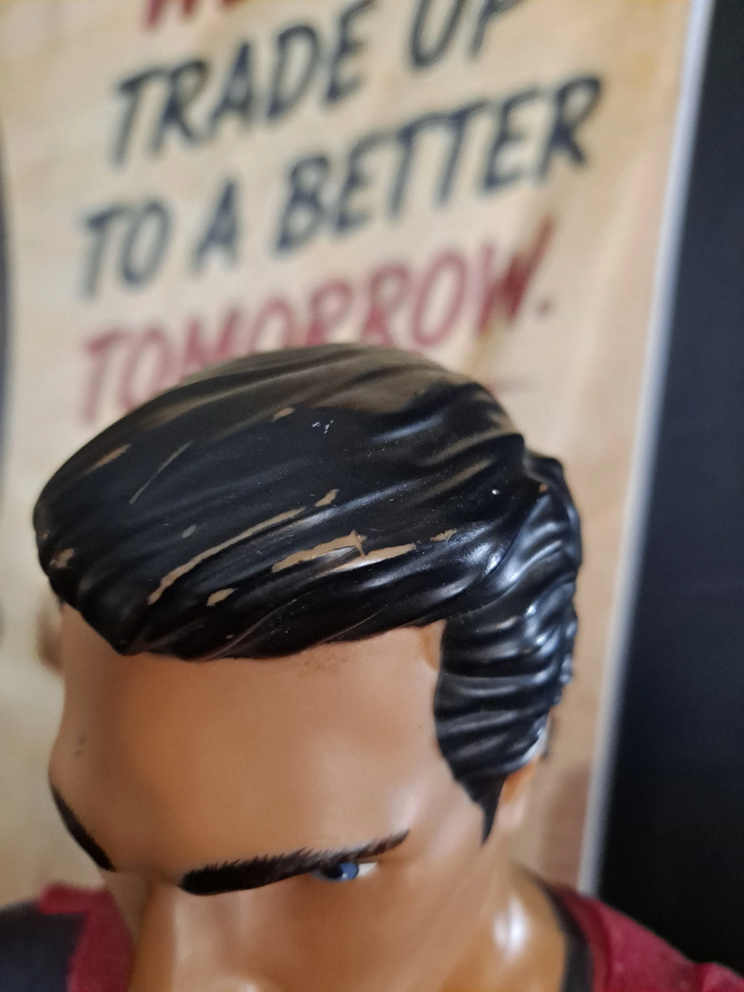 Close-up of a statue's face with black hair and blue eyes, with the background showing part of a sign with the words 'TRADE U' and 'TO A BETTER'.