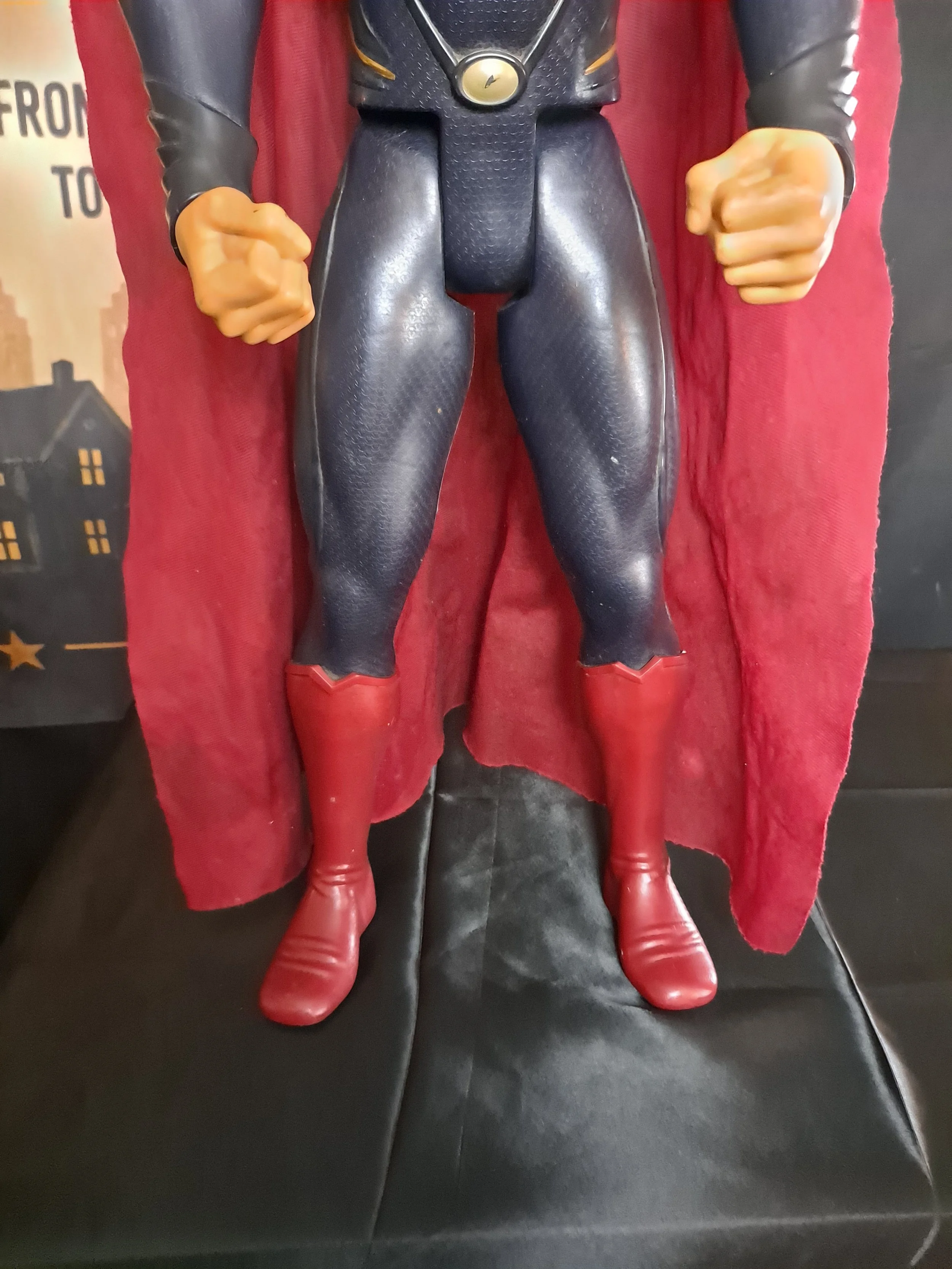 Close-up of a superhero figure dressed in a black and red costume with red boots, standing on a black surface with a red curtain behind.