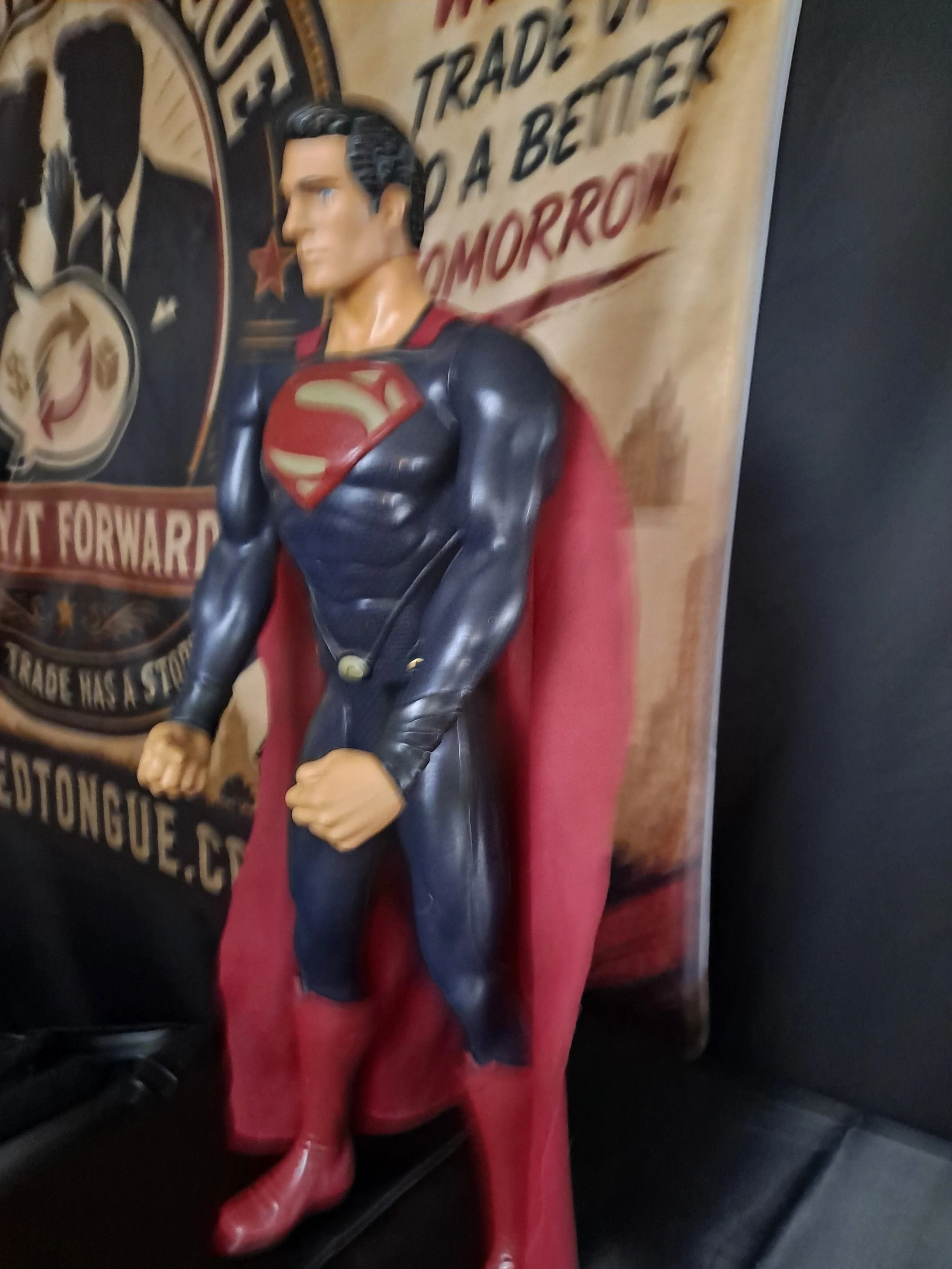 Close-up of a Superman action figure with a blurred background of a poster featuring Superman and text about trading.