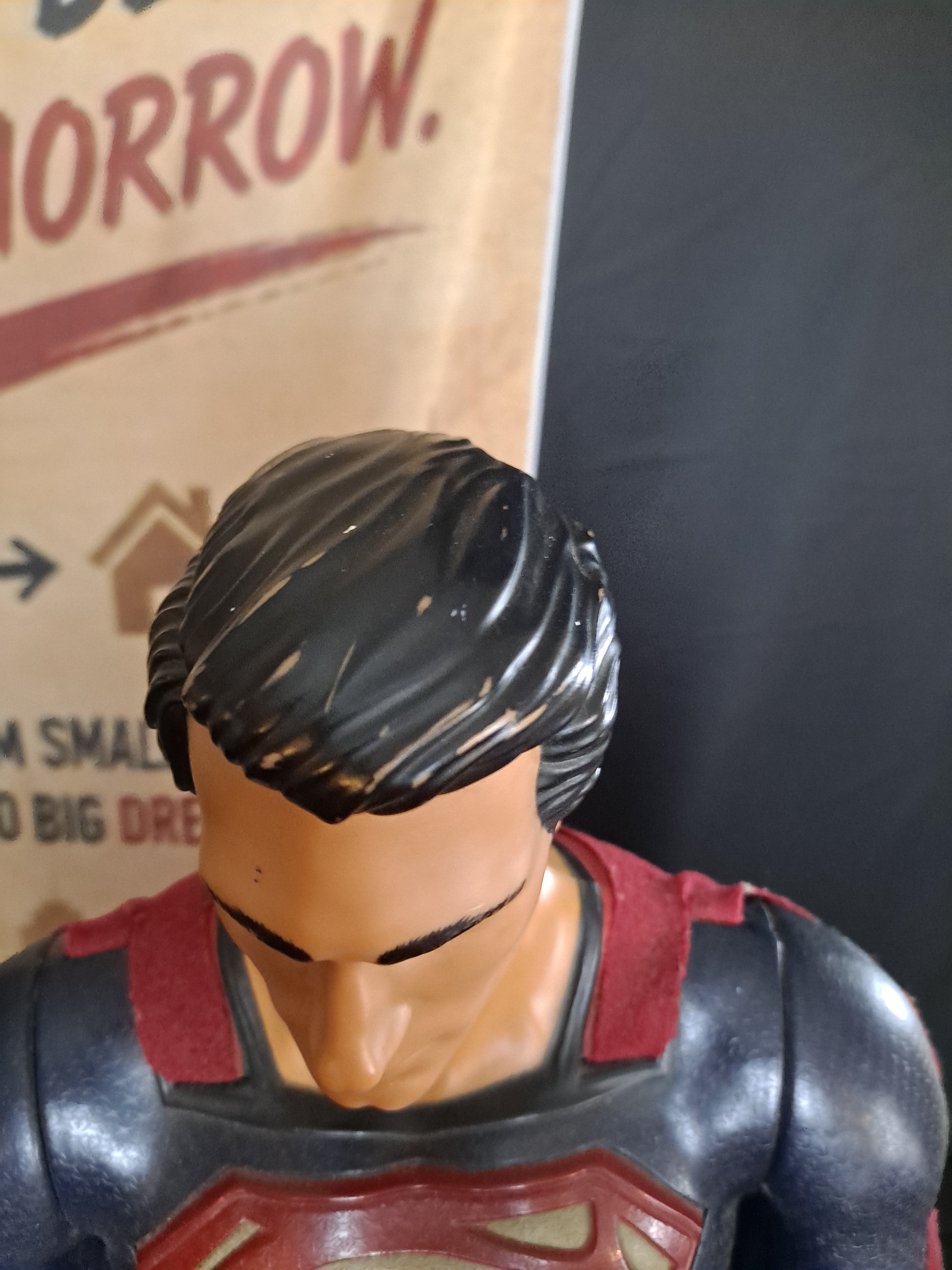 Close-up of a figurine resembling Superman with a red and blue costume and the Superman emblem on the chest. The figure has black hair with a curl and is wearing a black choker. The background includes a partially visible sign with the text 'TOMORROW' and some other words.