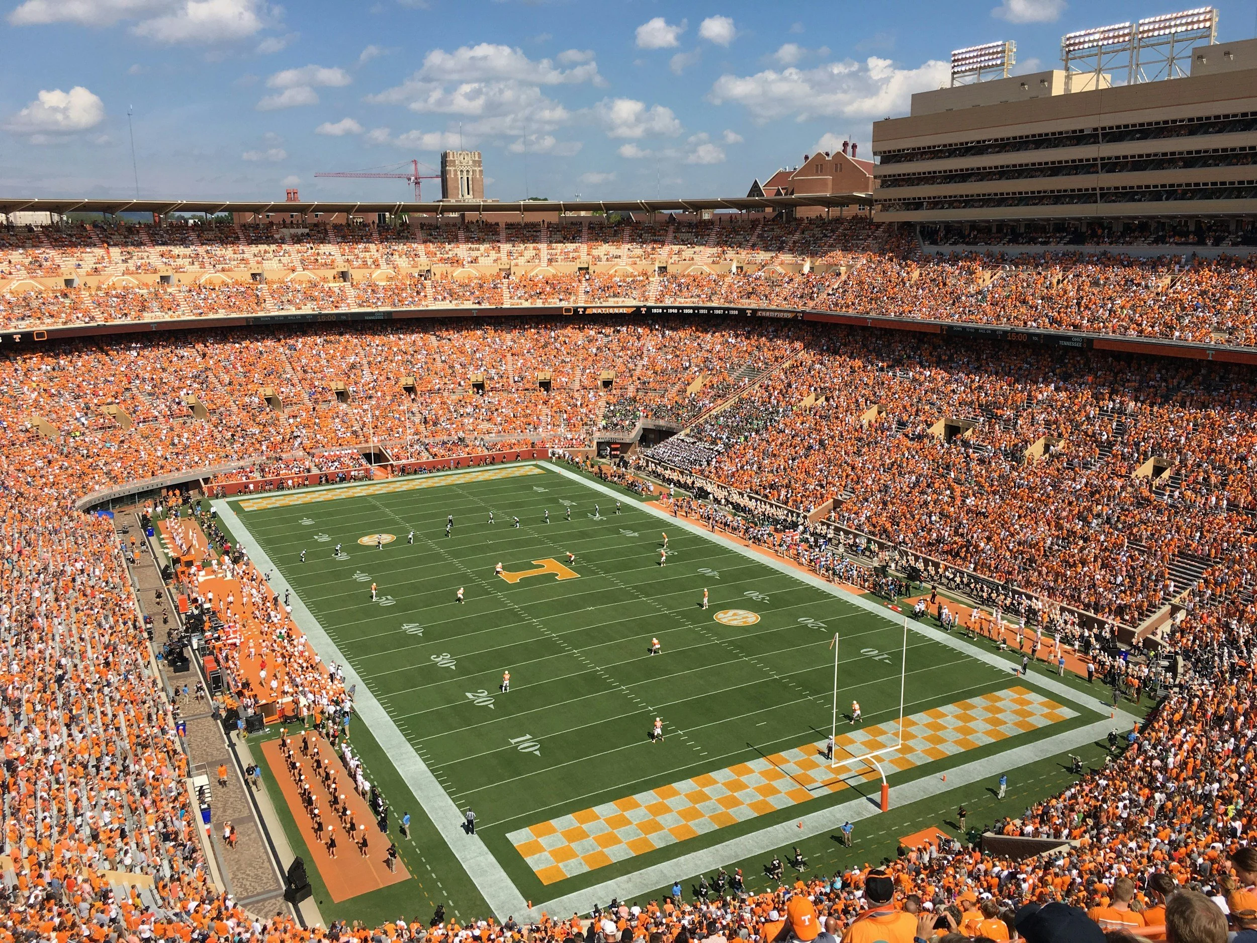 A large football stadium filled with fans wearing orange, with players on the field and a scoreboard showing the game is in the fourth quarter.