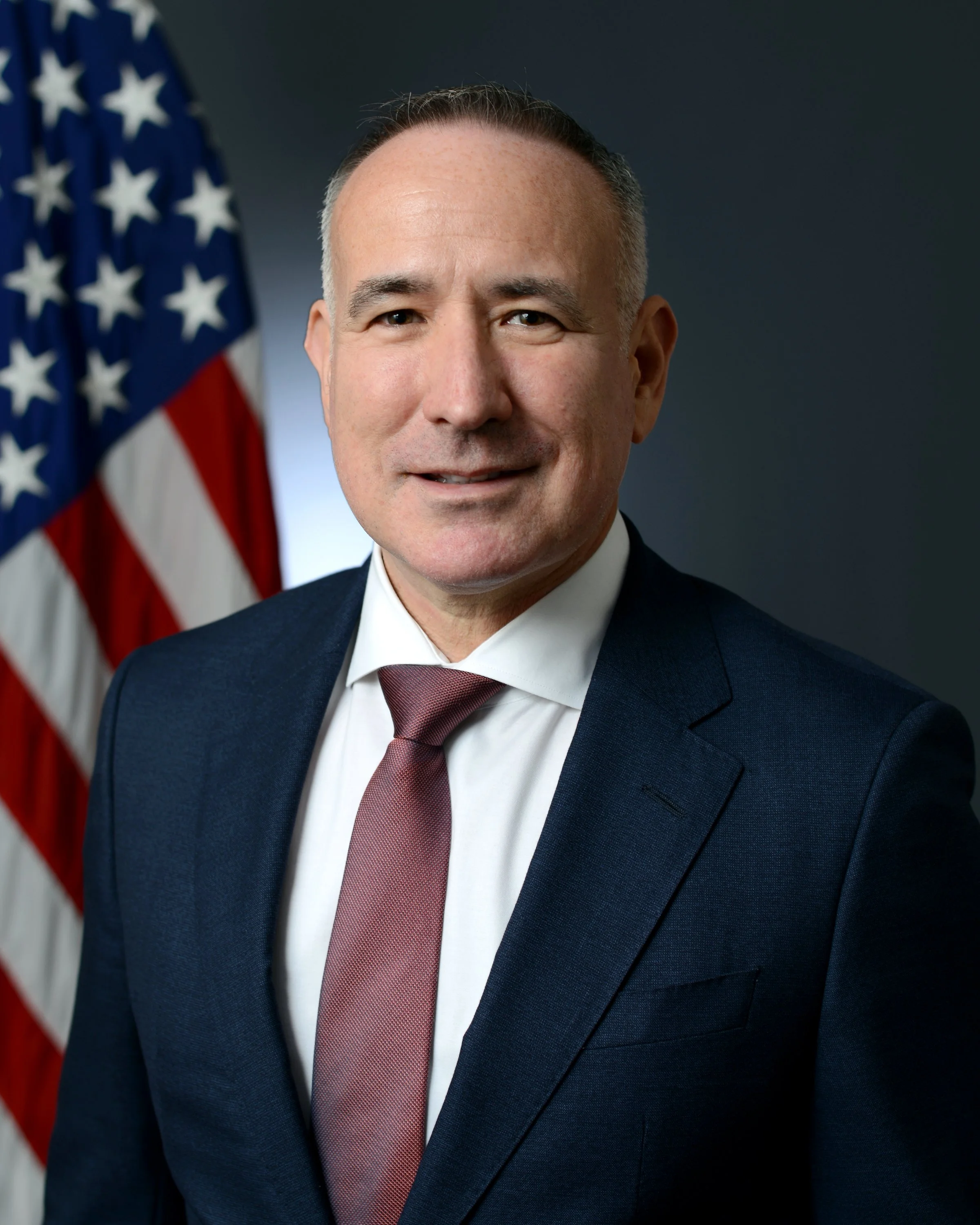Official portrait of Dr. Albert “Jim” Marckwardt in a dark suit and maroon tie, standing beside the American flag.