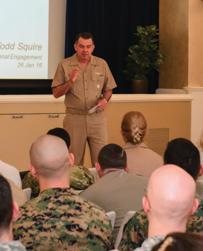 A military officer giving a presentation to soldiers seated in a room with a projector screen, a potted plant, and a blue curtain.