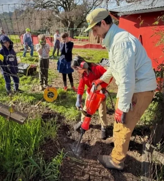 A group of children and an adult working together in a garden, with the adult using a power drill to dig in the soil while others observe nearby.
