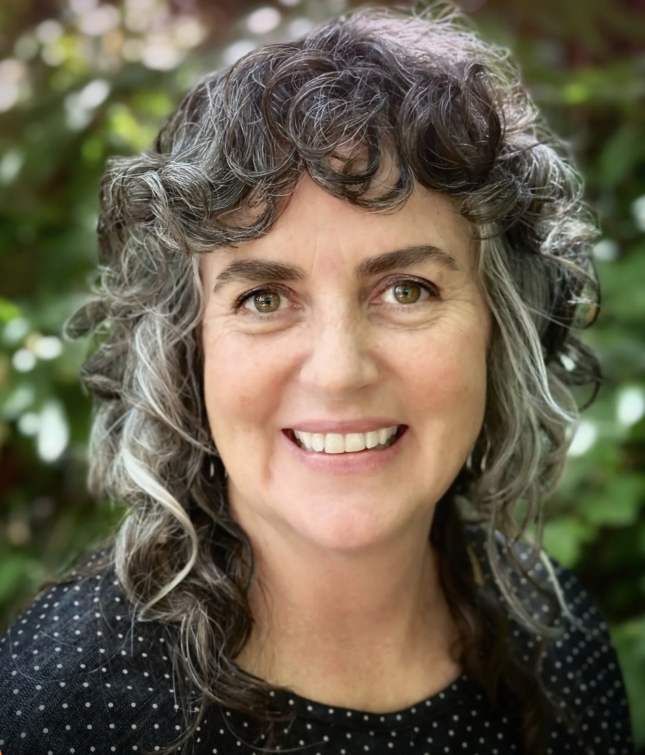 Close-up of a woman with curly gray hair smiling outdoors with a background of green foliage.