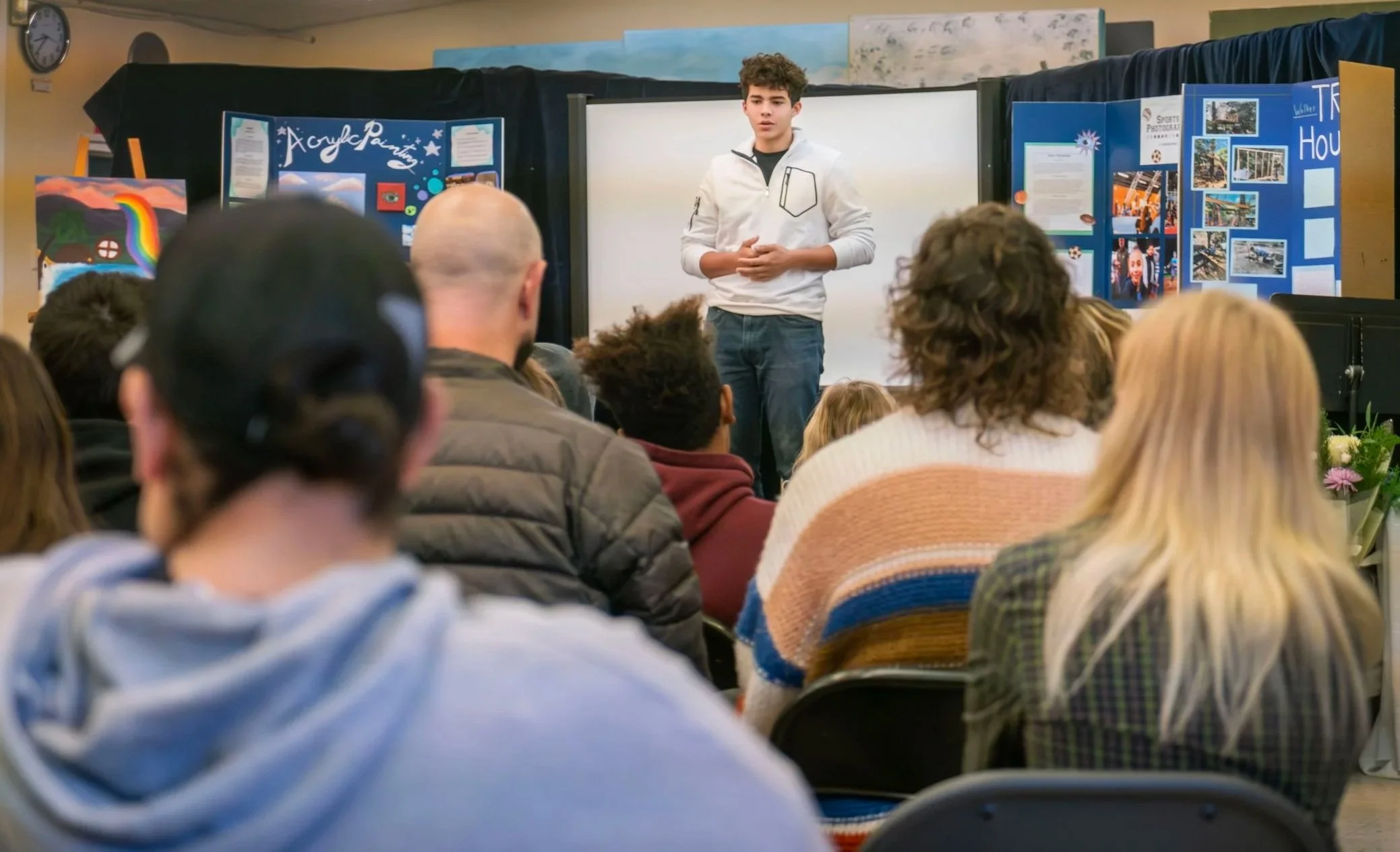 A young man giving a presentation in front of an audience, with displayed posters and artwork on the background walls.
