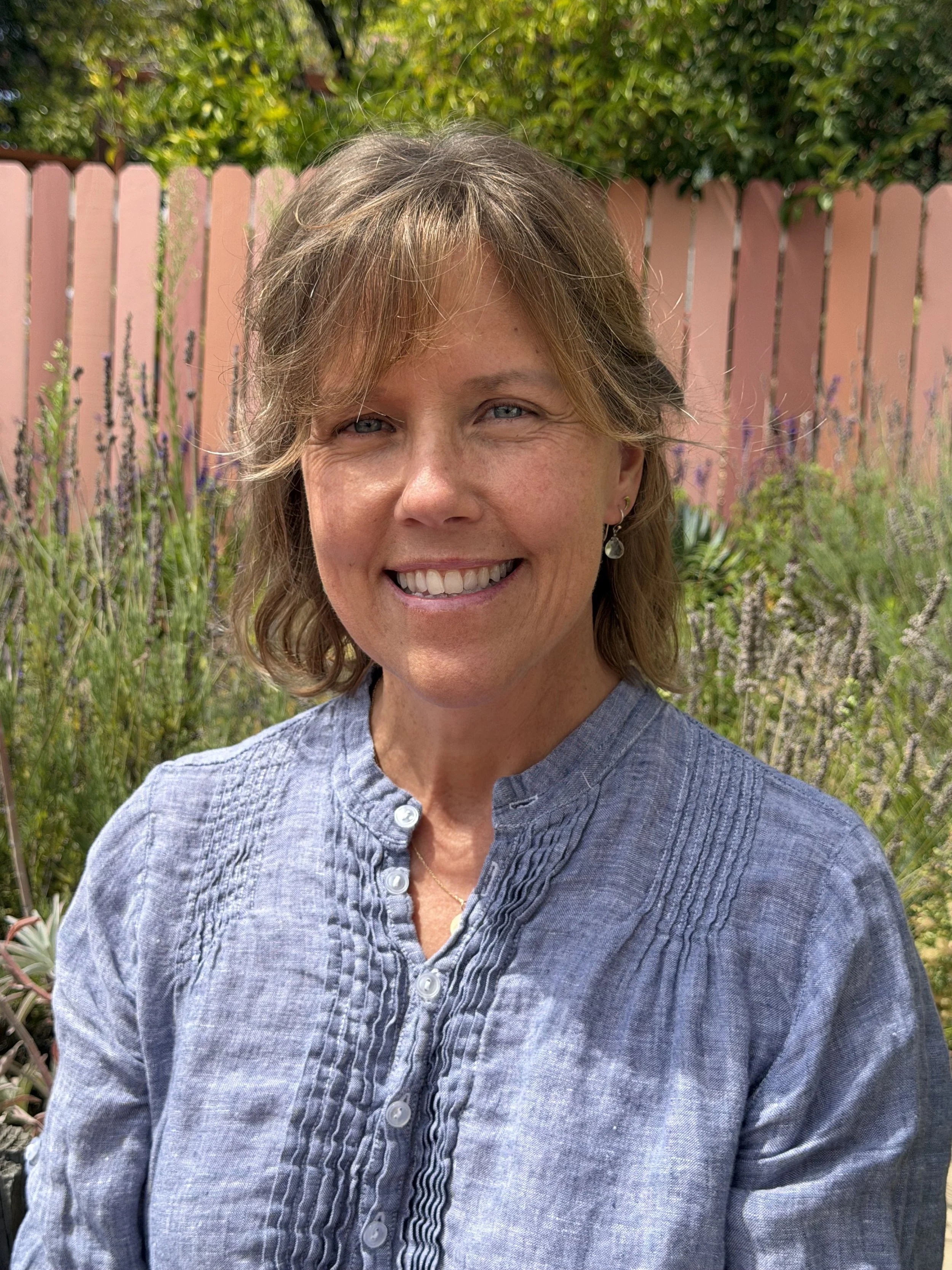 A smiling woman with shoulder-length light brown hair, wearing a blue button-up shirt, standing outdoors with a pink wooden fence and green foliage in the background.