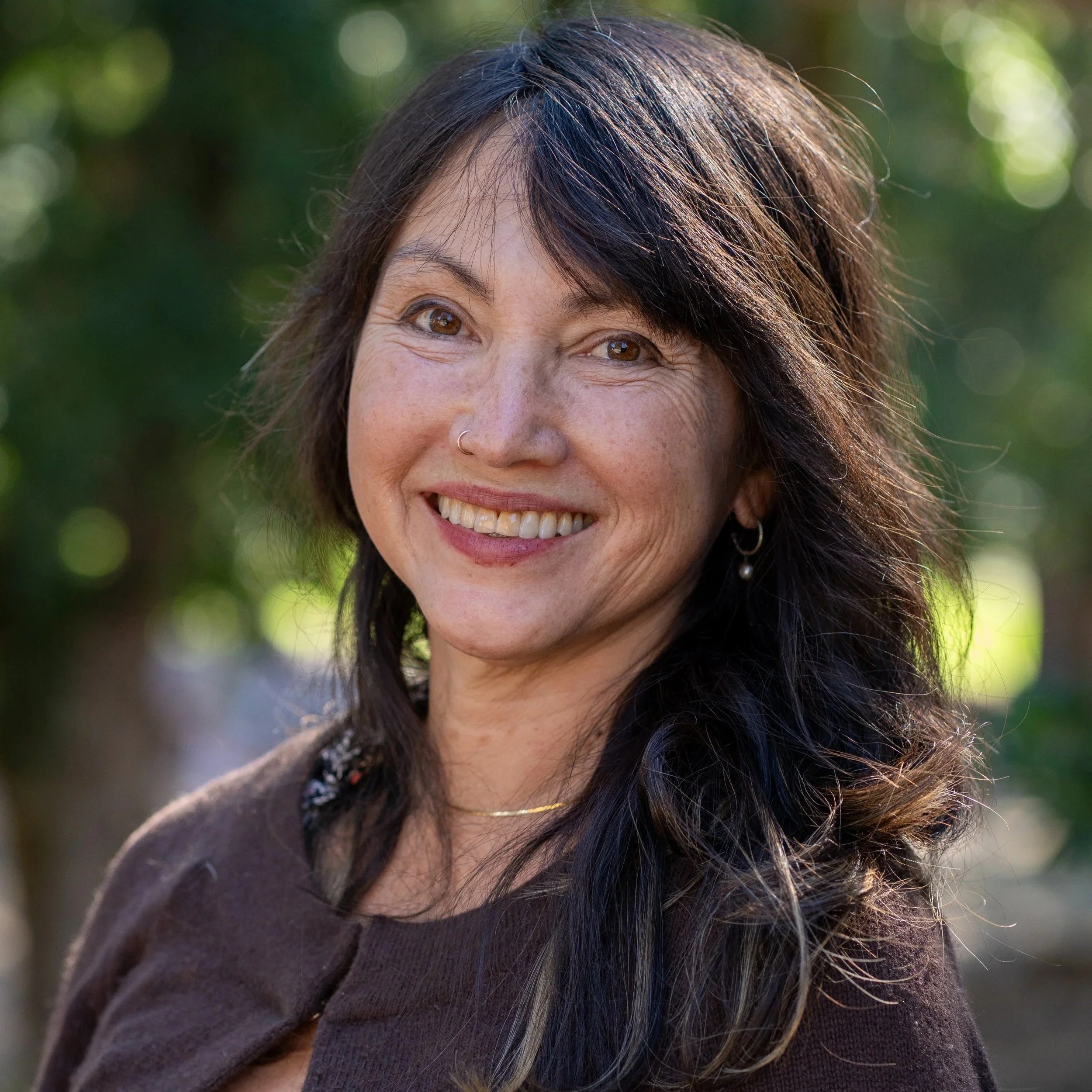 Close-up of a middle-aged woman with dark brown hair and brown eyes, smiling outdoors with a blurred green background.