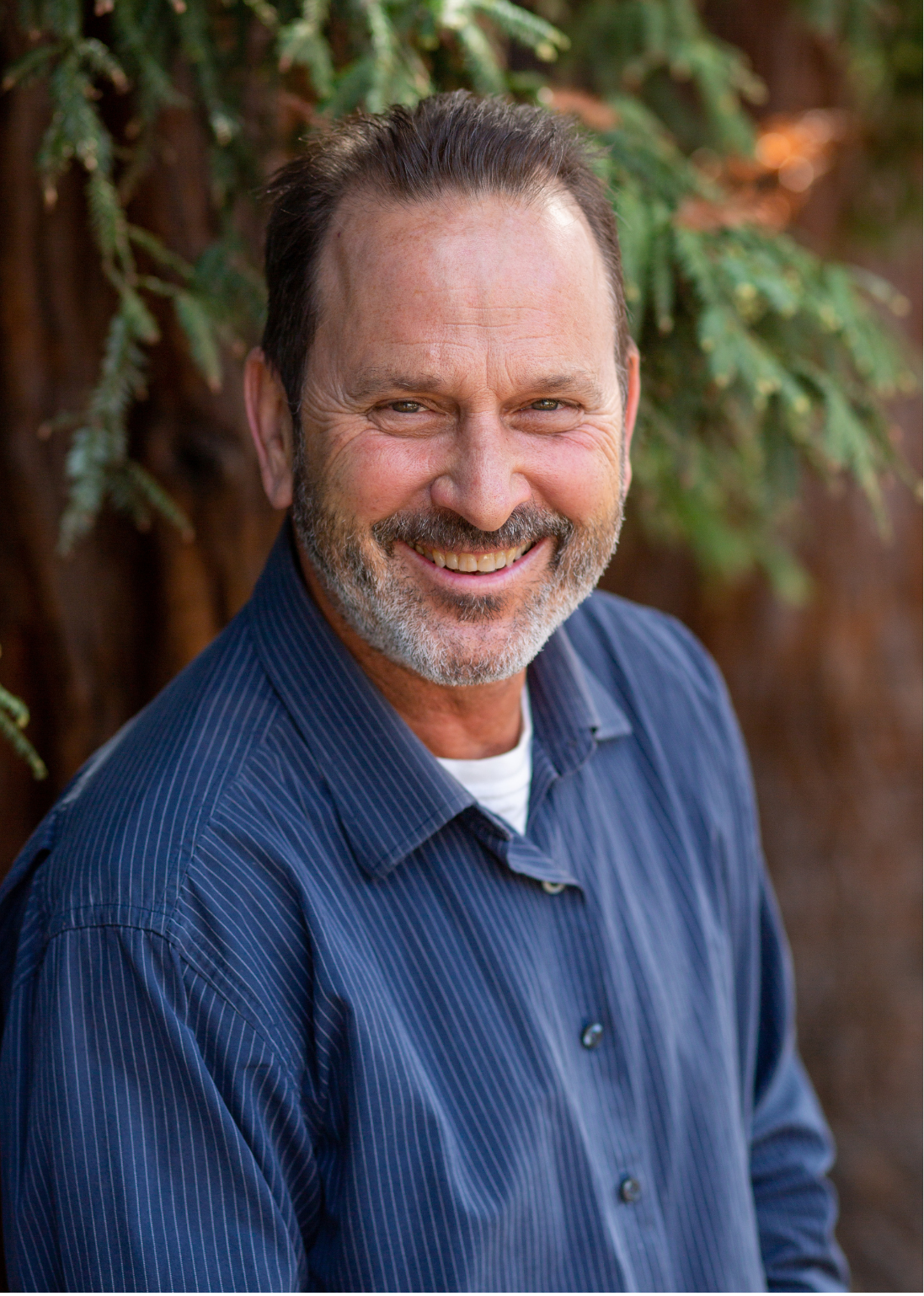 A middle-aged man with a gray beard and short dark hair smiling outdoors in front of a tree with green foliage.