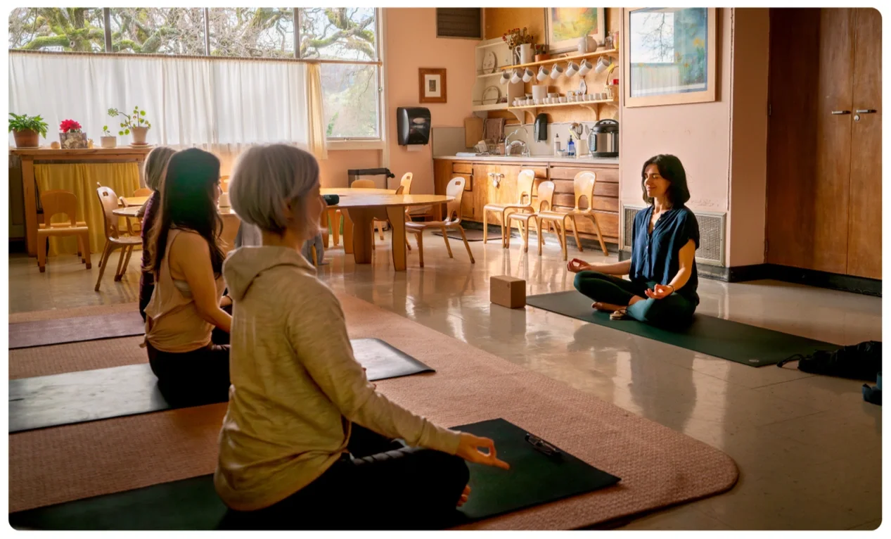 A group of women and girls doing yoga meditation in a cozy kitchen area with natural light, wooden furniture, and open shelves.