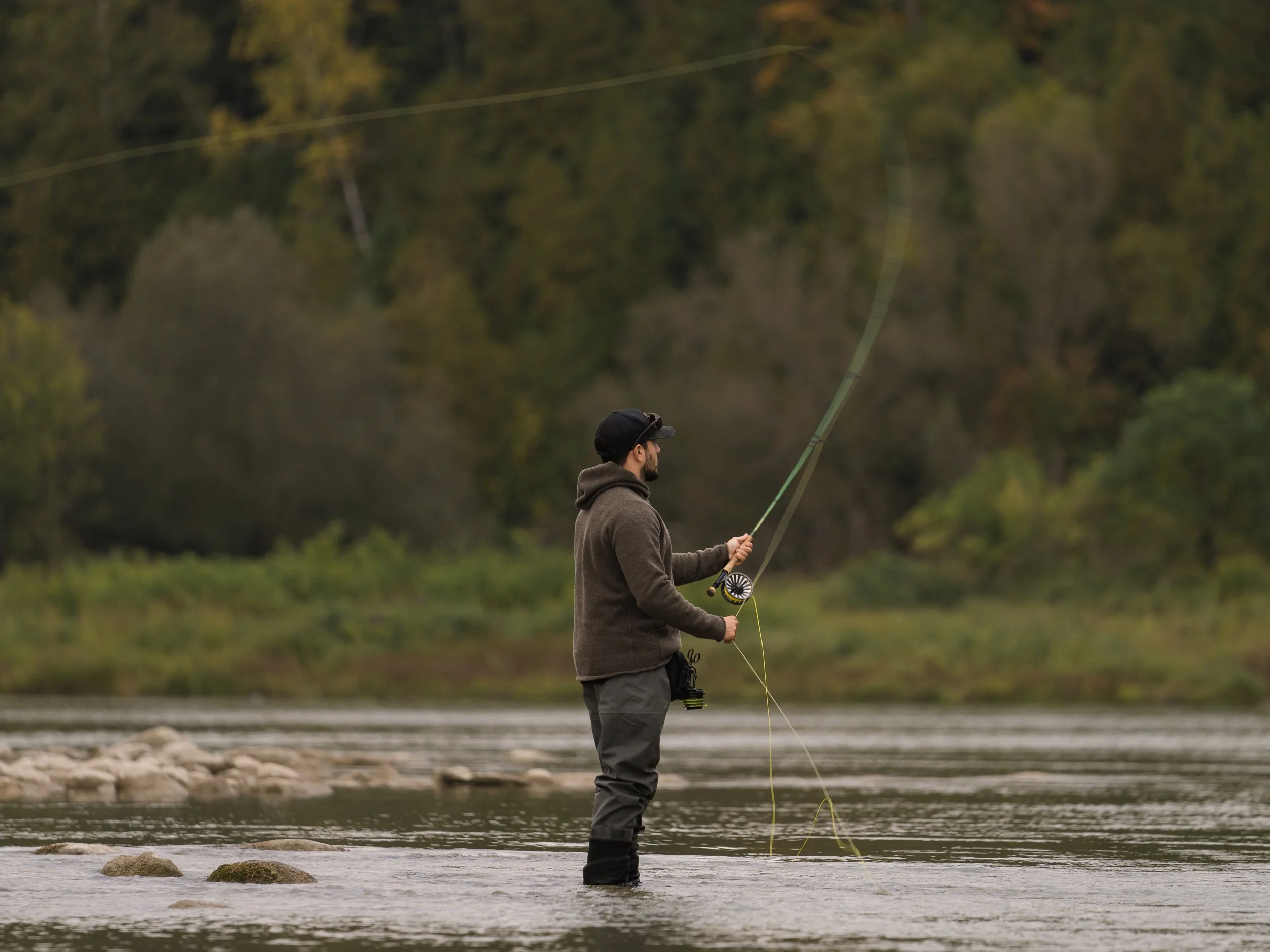 Fly angler in a hooded jacket casting while wading a calm, forest-lined river