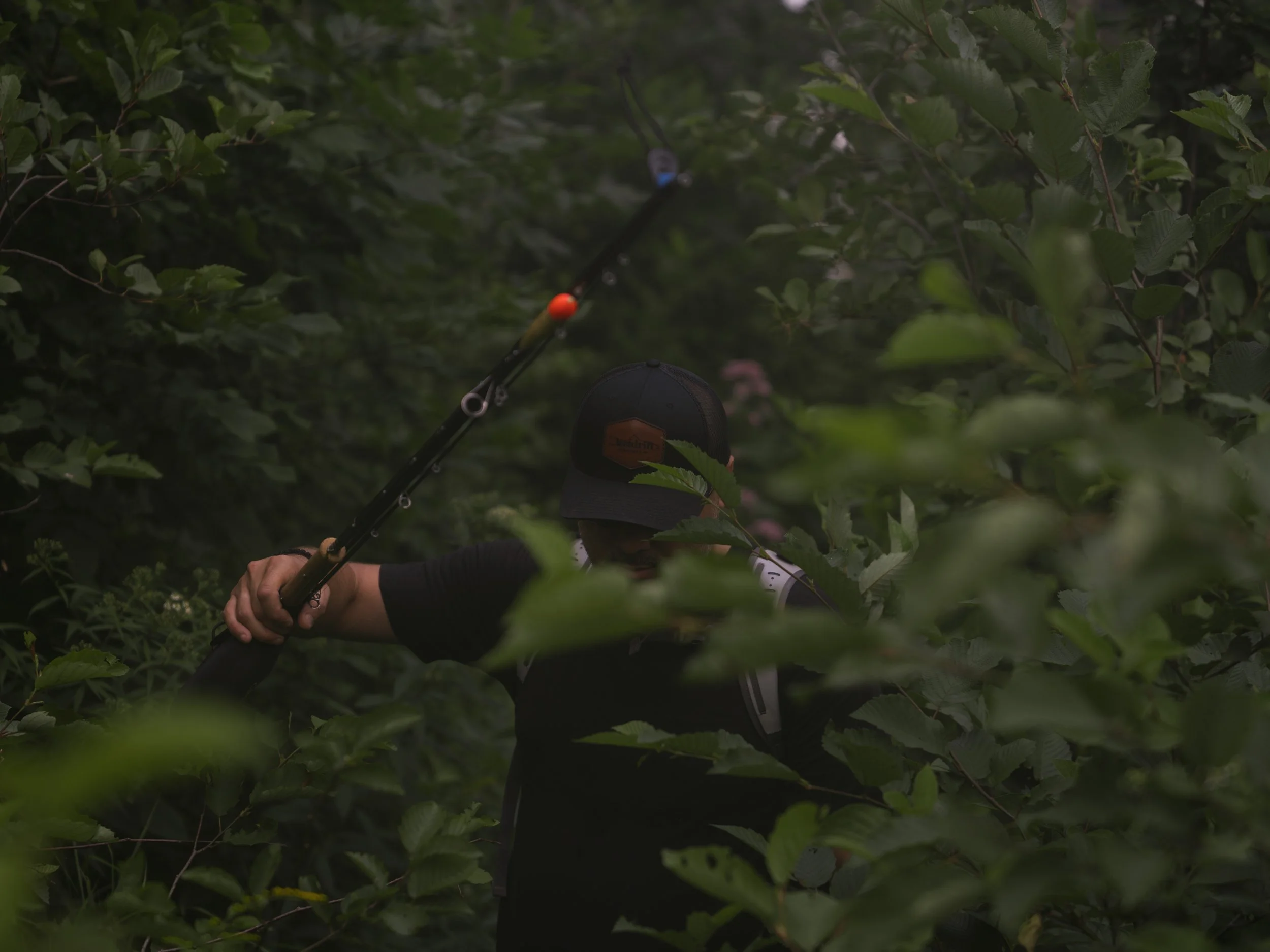 Angler in a cap pushing through thick green brush, holding a rod