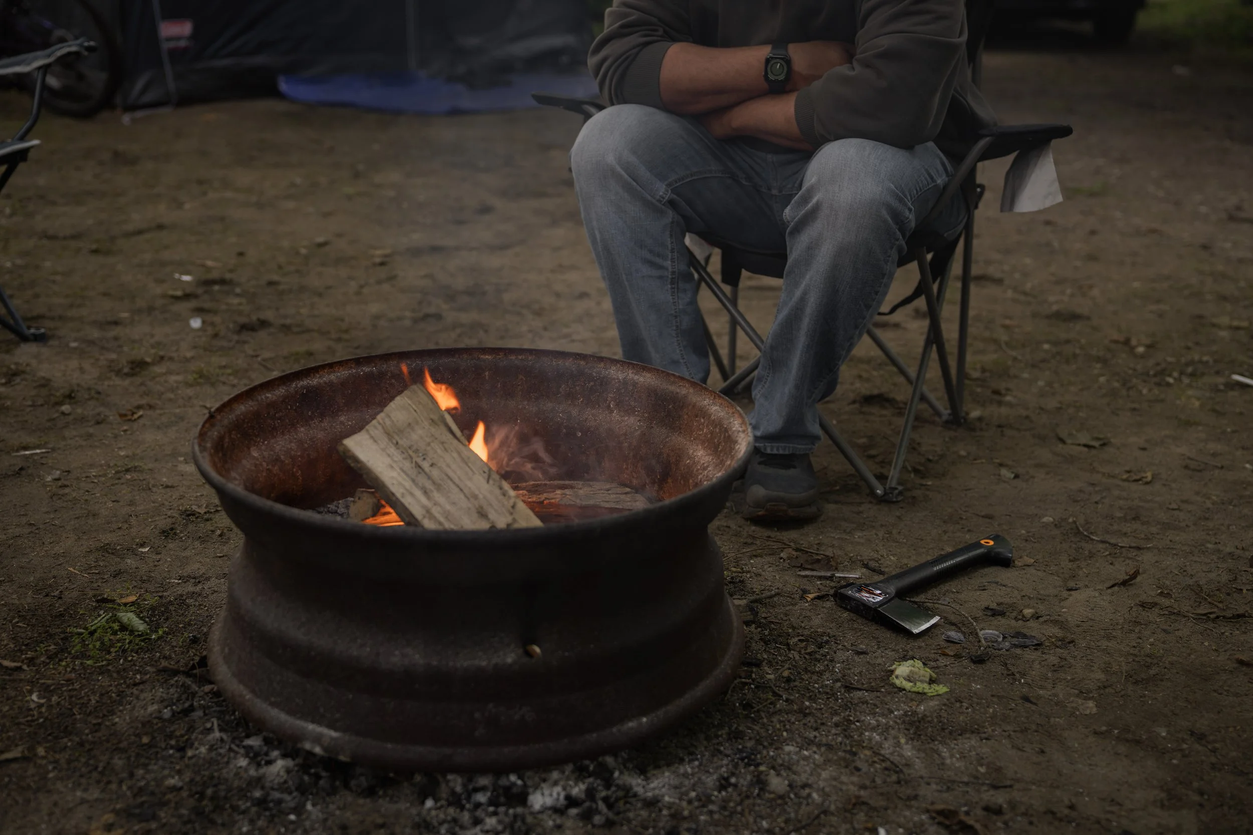 Person relaxing in a camp chair beside a steel fire pit with a burning log