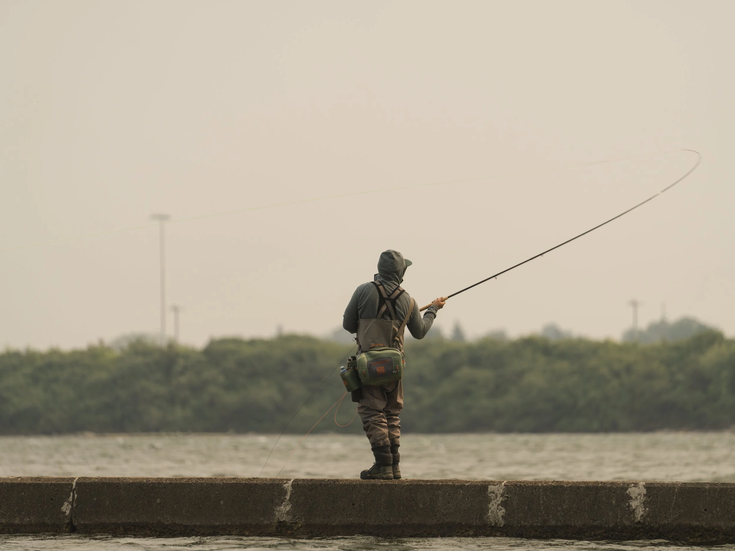Fly angler in Simms waders casting from a concrete breakwater into a river under hazy grey skies