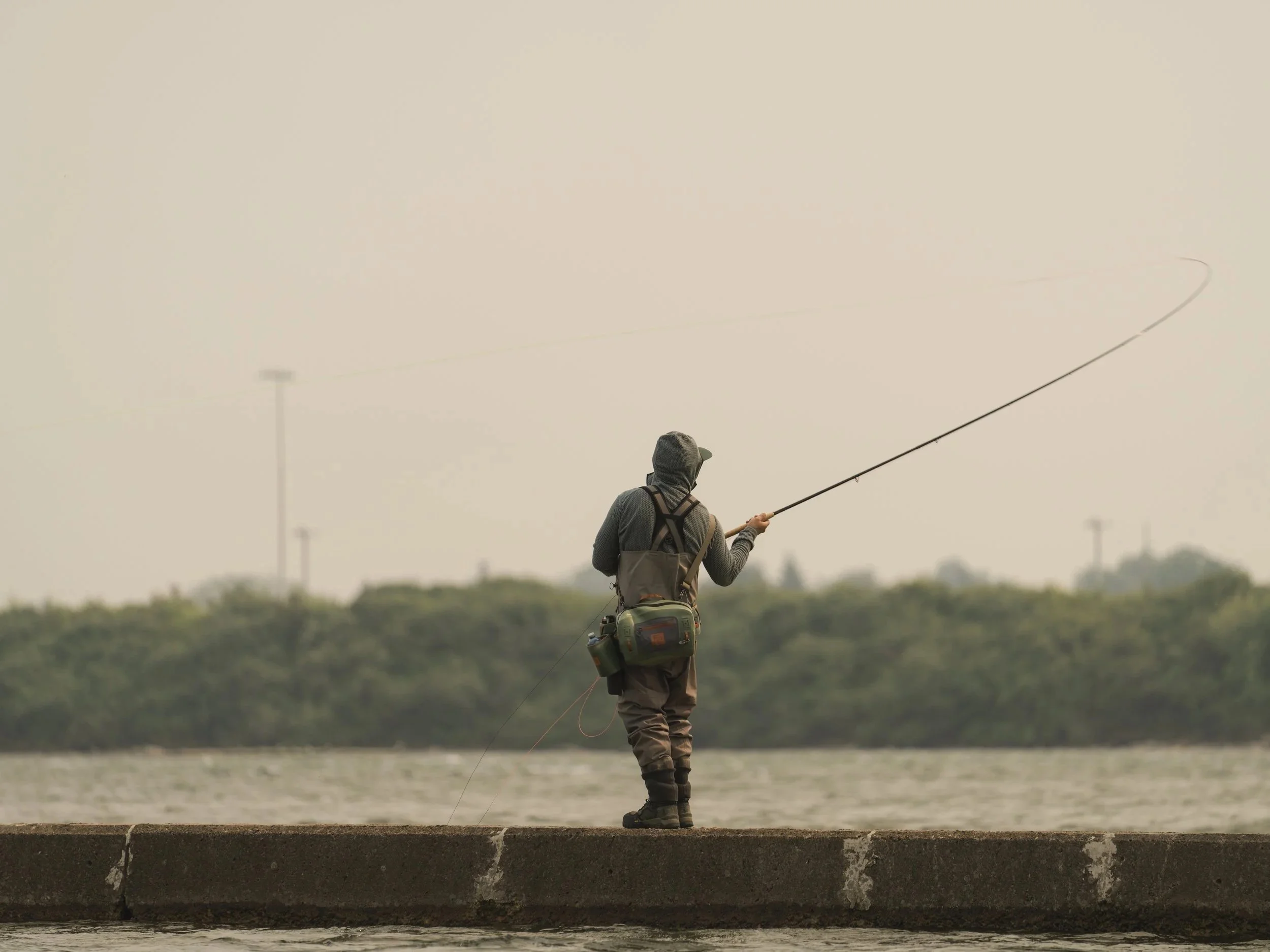 Fly angler in Simms waders casting from a concrete breakwater into a river under hazy grey skies