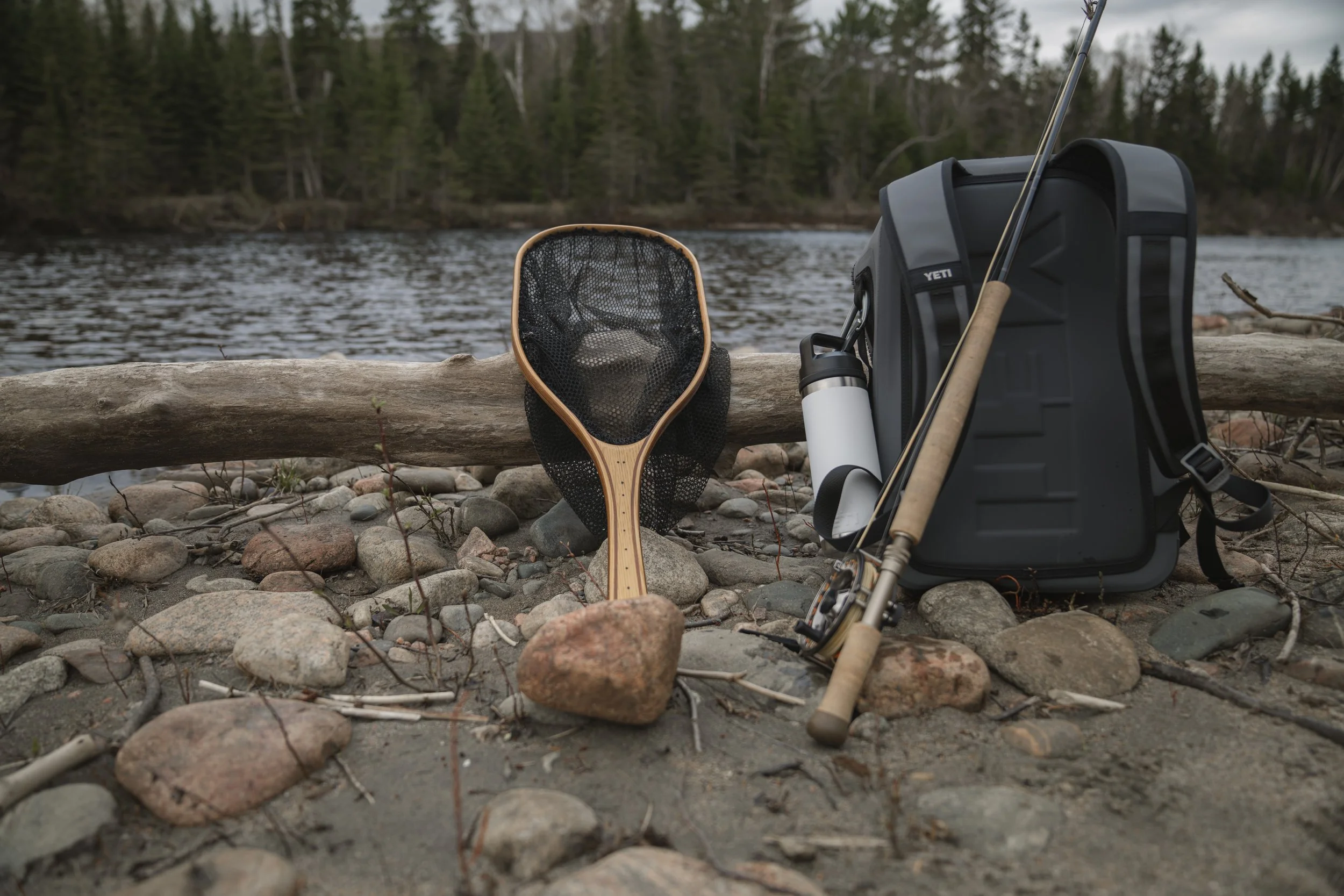 Cedar landing net, Yeti backpack and Sage fly rod staged on a rocky riverbank beside a fallen log