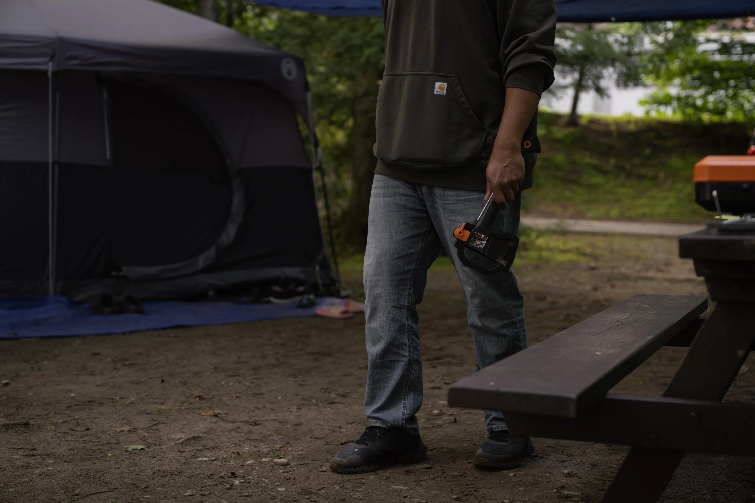 Camper carrying a Fiskar's axe past a Coleman tent and picnic table at a wooded campsite