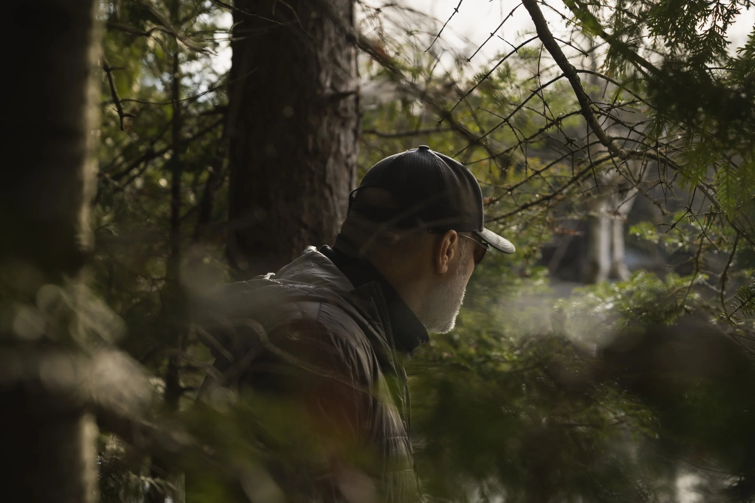 Person in a cap standing quietly in a dark forest, foliage blurred across the foreground
