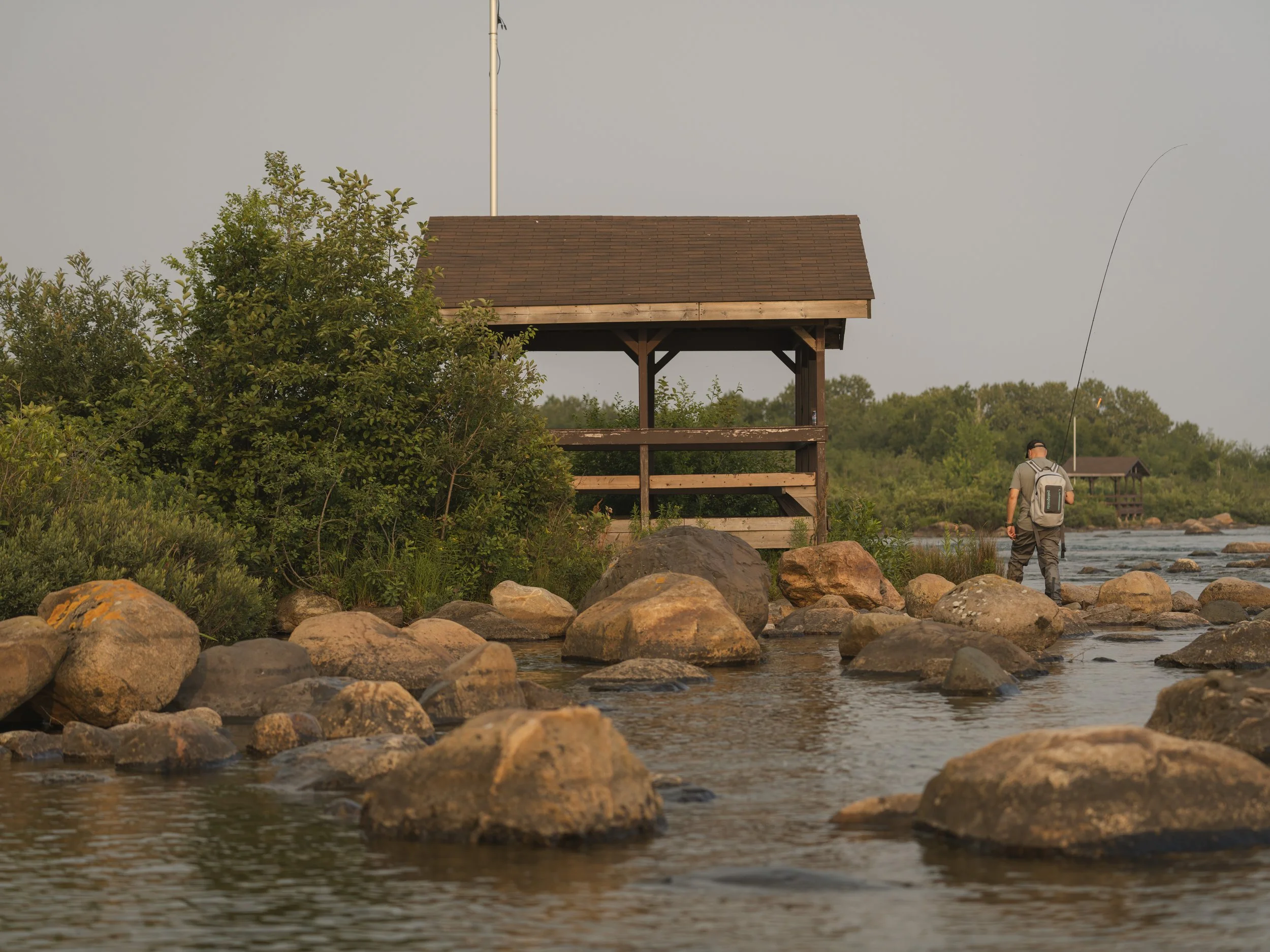 Wooden riverside shelter above boulder-strewn shallows with an angler wading nearby