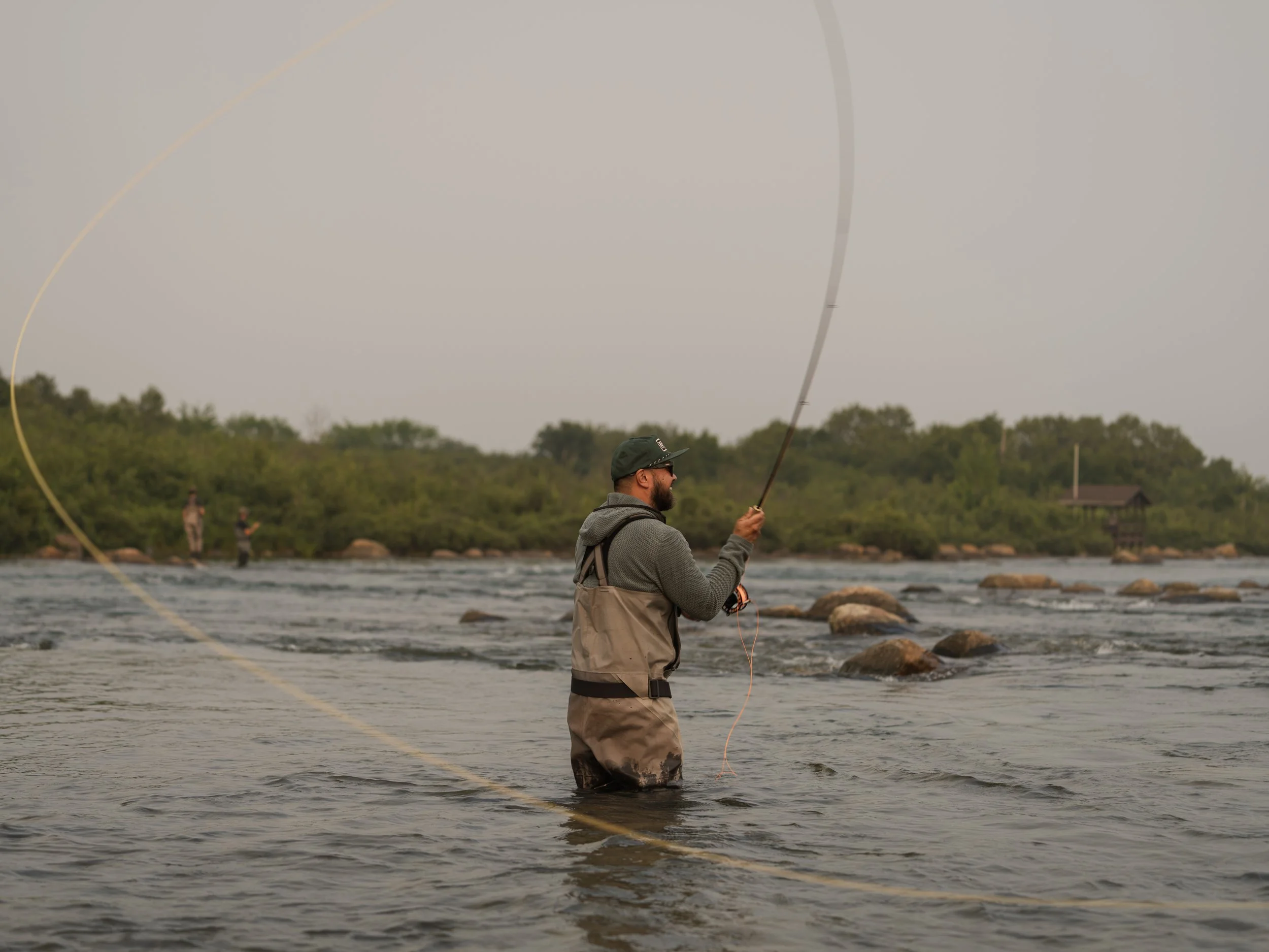 Angler in Simms chest waders casting a wide line loop, a second angler fishing downstream