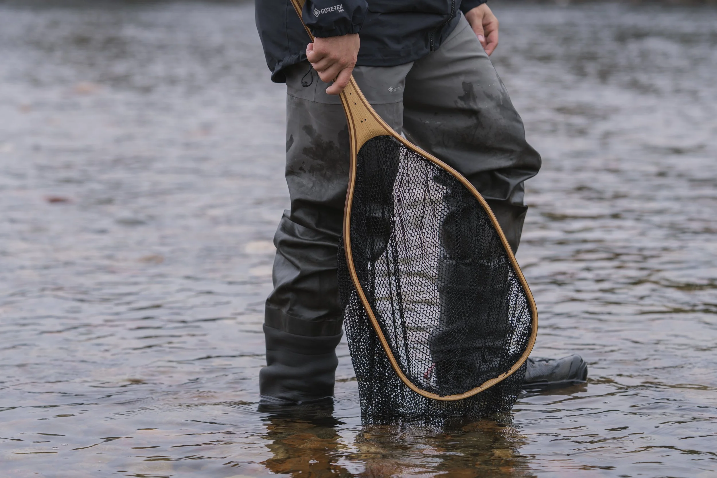 Angler in  Simms waders standing in the shallows with a wooden Cedar landing net