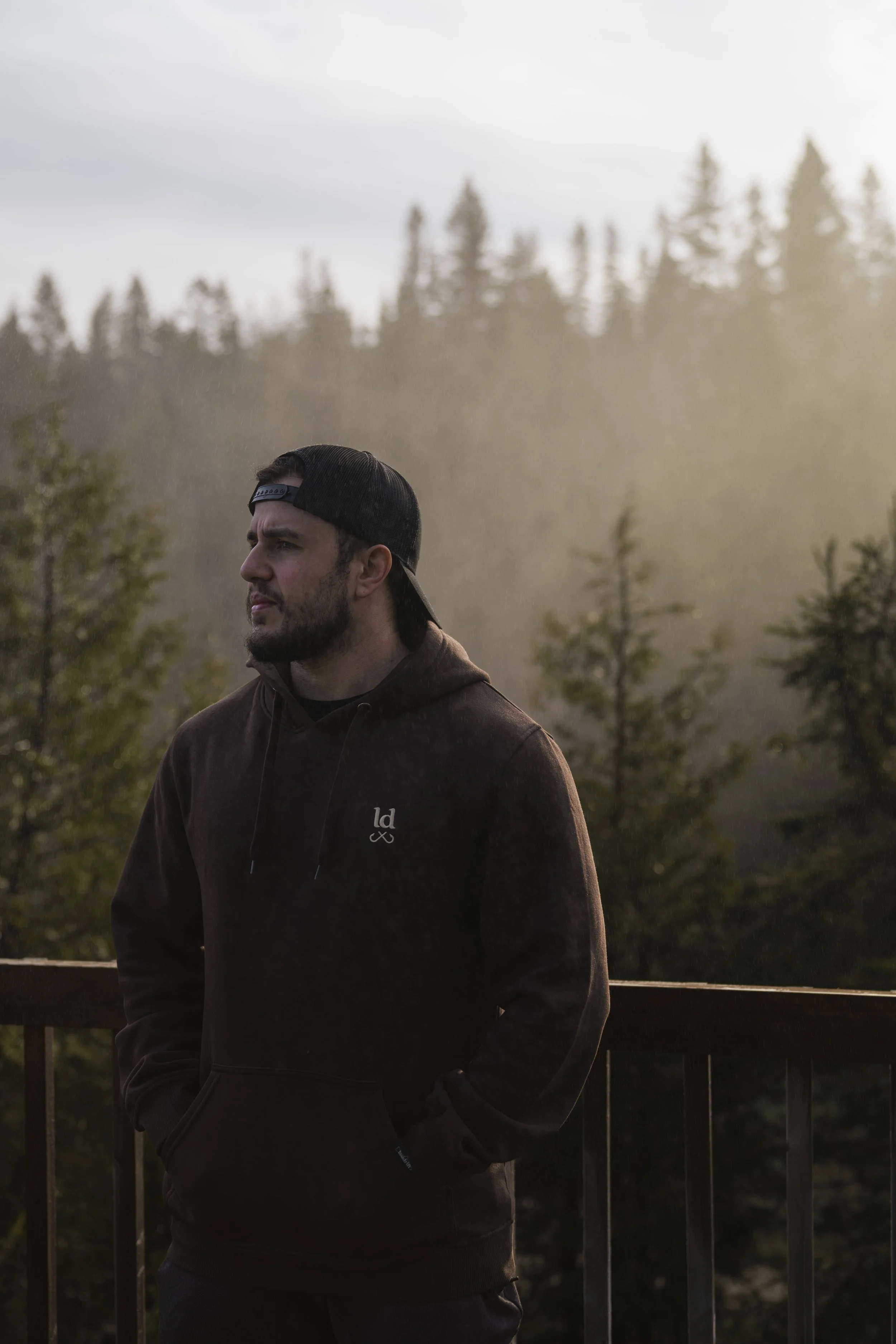 Man in a backwards cap and a Lastdrift hoodie at a railing, gazing over a misty evergreen forest at golden hour