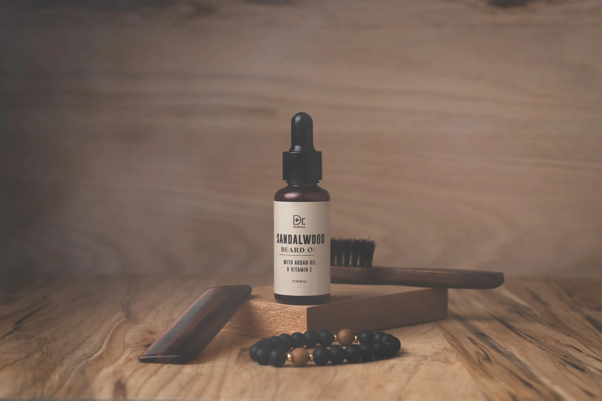 Sandalwood beard oil bottle styled with a beard brush, Kent comb and beaded bracelet on a warm wood surface