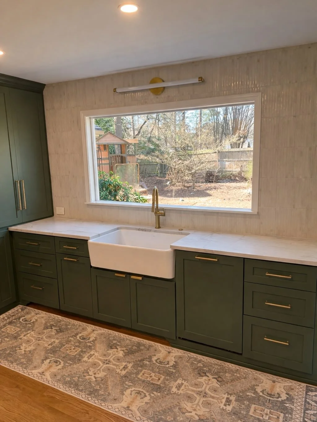 Kitchen with green cabinets, white countertop, farmhouse sink, large window overlooking a backyard with trees, and a patterned rug on wooden floor.